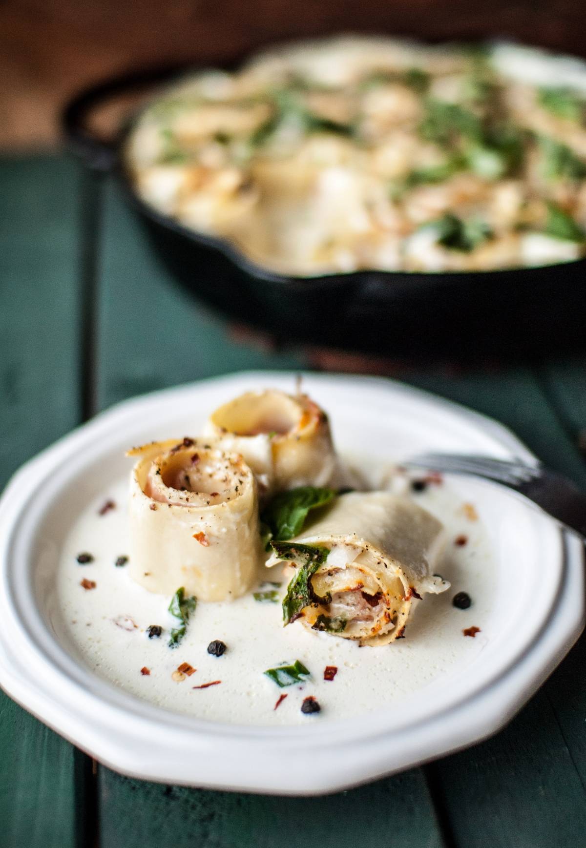Two Lasagna Roll Ups sit on a white plate with creamy sauce and herbs, while more pasta pinwheels bake in a skillet behind.