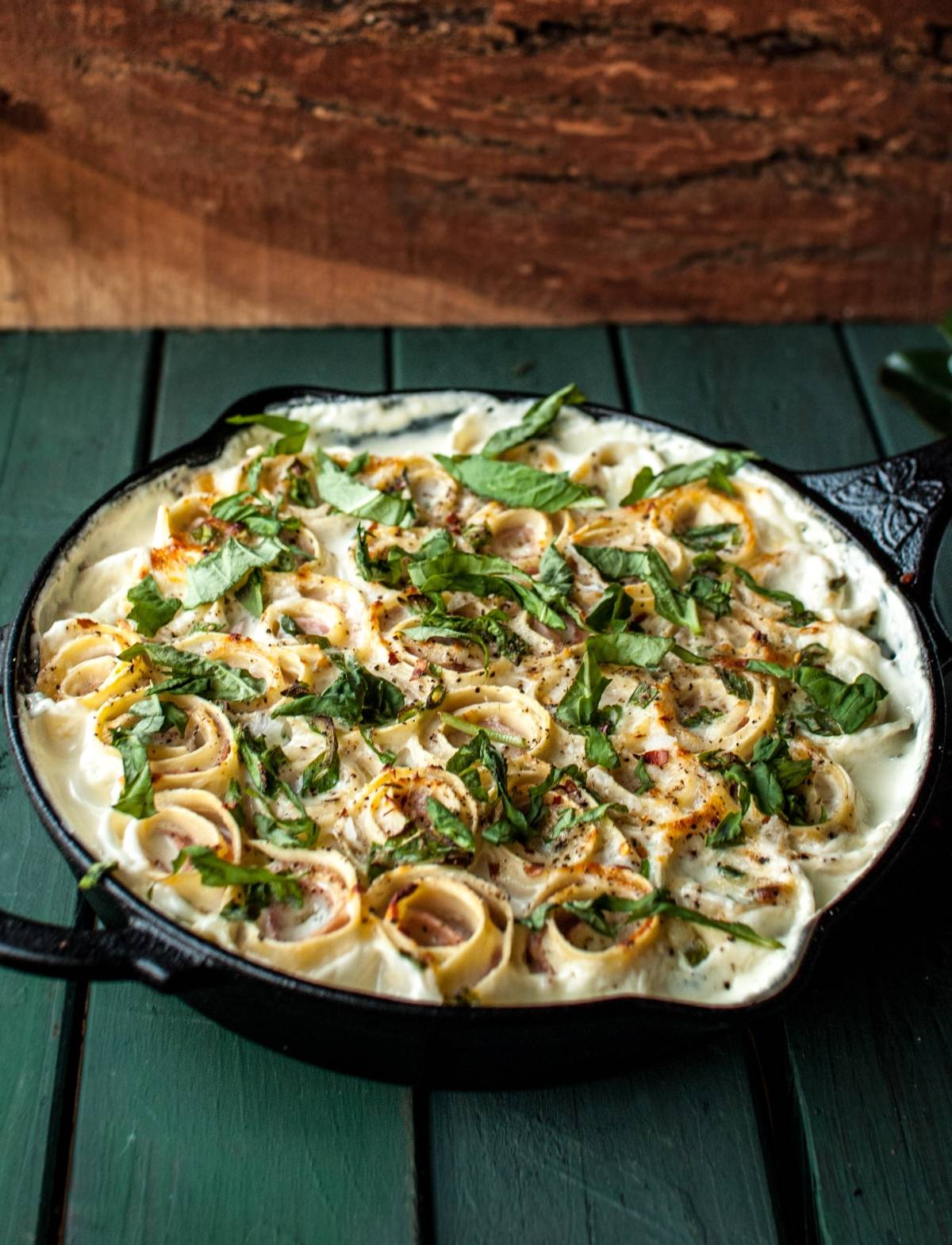A skillet of creamy baked pasta roses topped with fresh herbs, sitting on a green wooden surface.