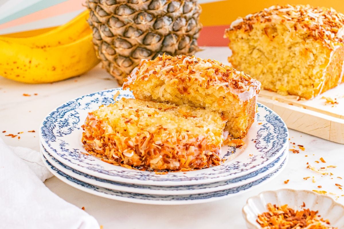 Sliced Hawaiian banana bread with pineapple and coconut on a decorative plate, with bananas and a pineapple in the background.