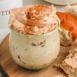A jar of homemade cinnamon butter sits on a wooden board, surrounded by sliced bread rolls and a butter container.