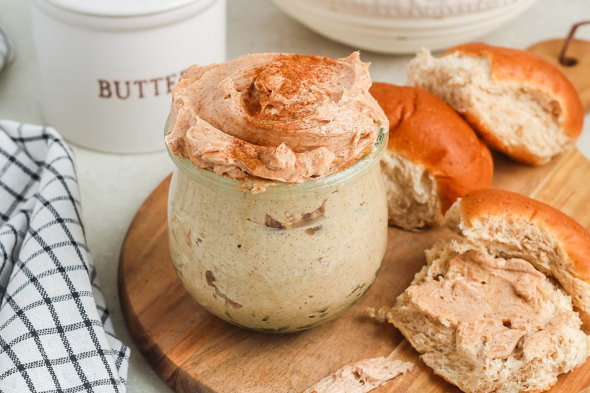 A jar of homemade cinnamon butter sits beside rolls and a checkered napkin on a wooden board.