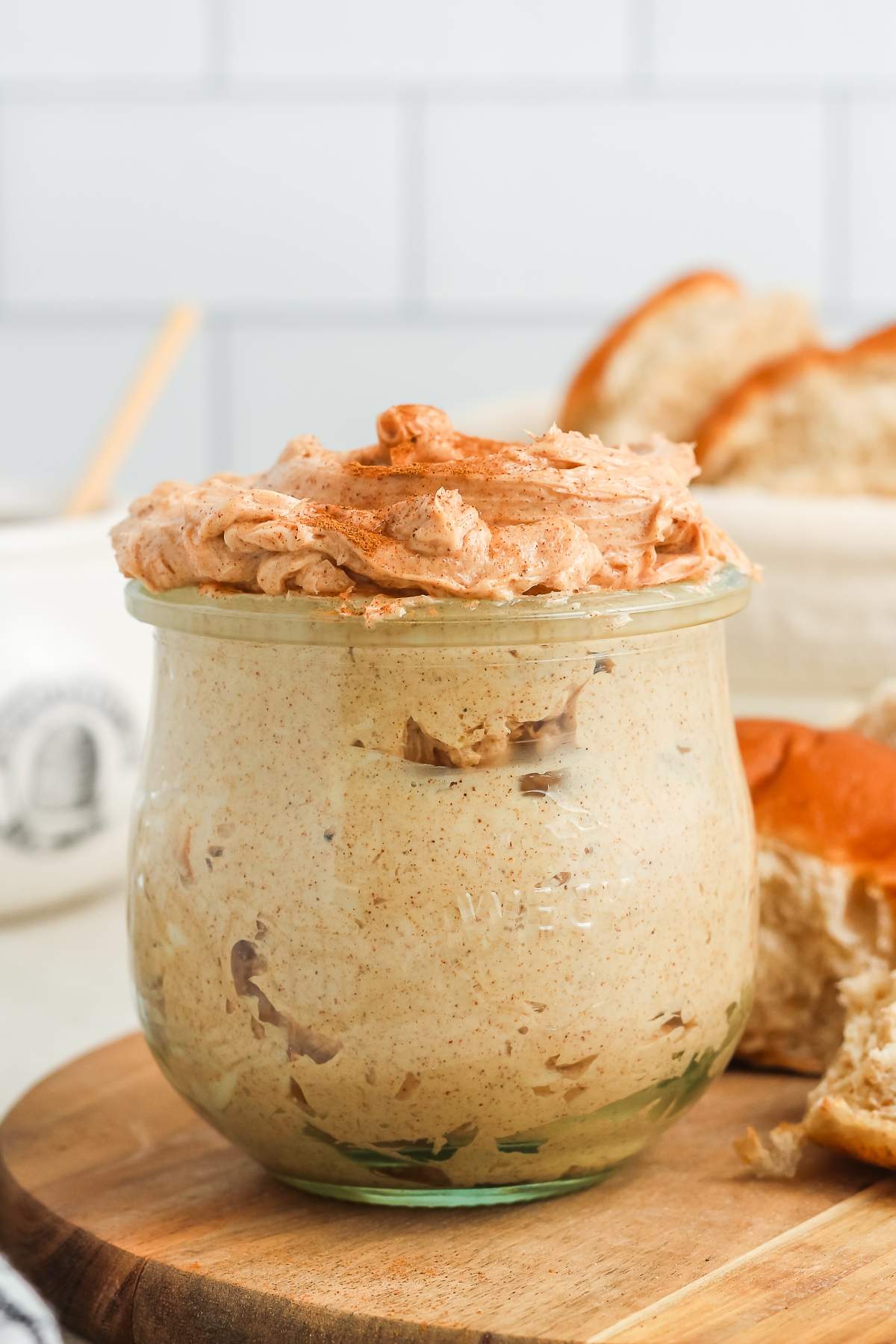 A glass jar filled with whipped cinnamon butter, placed on a wooden board with bread in the background.