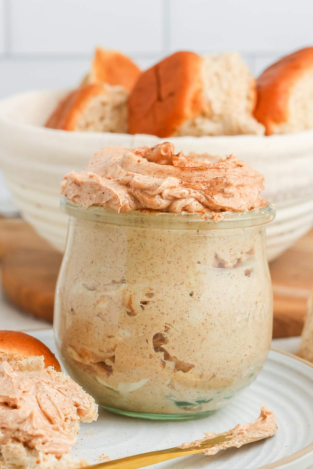 A jar of whipped cinnamon butter sits in front of a bowl of sliced bread rolls.