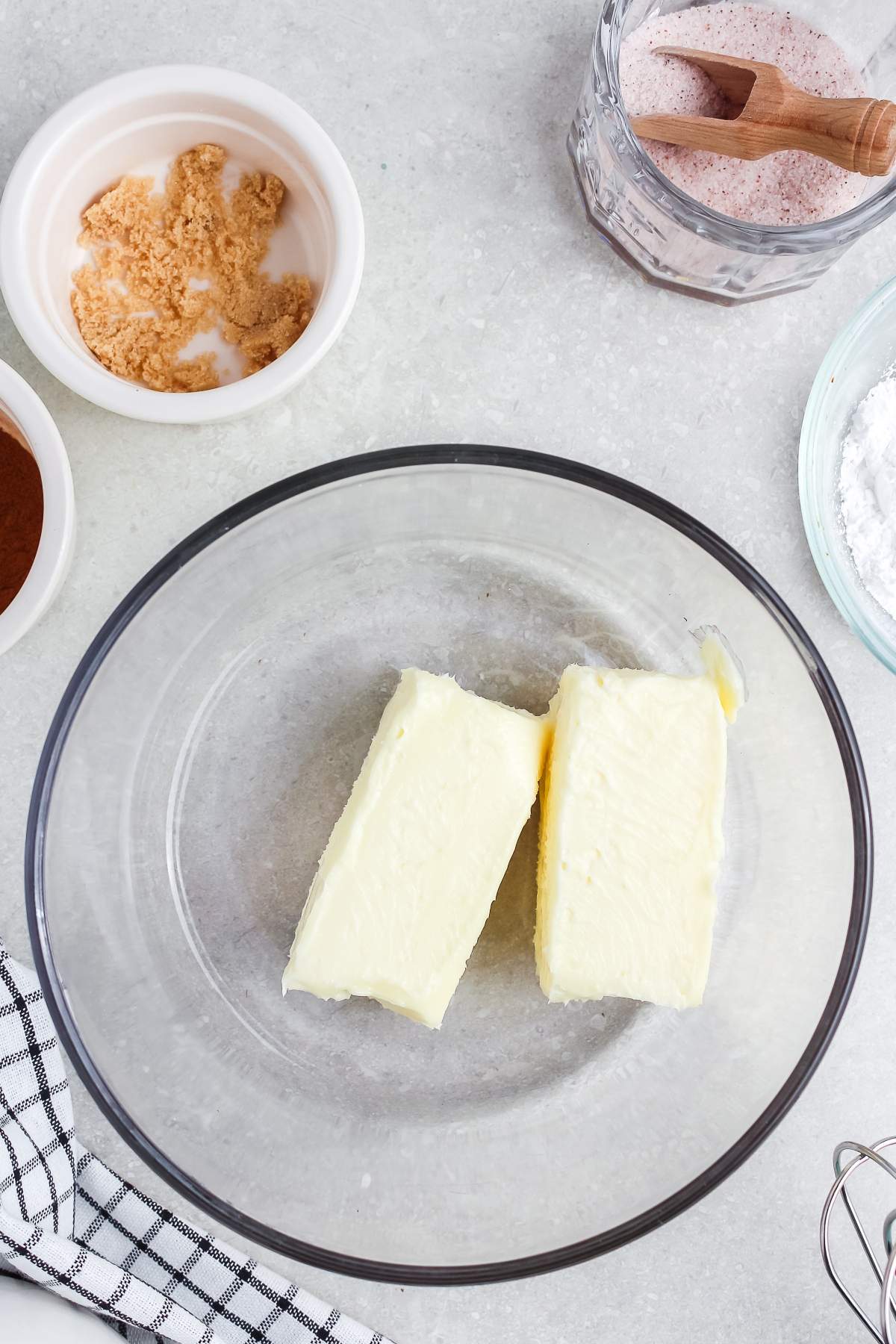 Two sticks of butter in a glass bowl, surrounded by bowls of sugar, salt, and other baking ingredients.