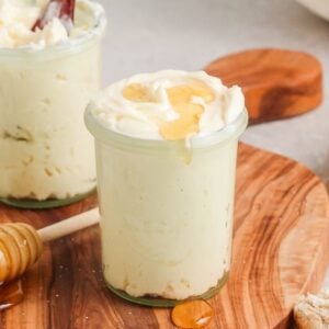 Two small jars of homemade honey butter on a wooden board, one topped with honey, sit next to a honey dipper and fresh bread.