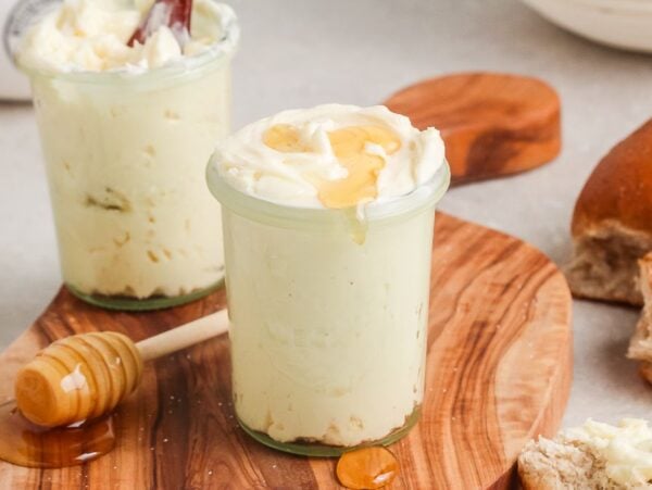 Two small jars of homemade honey butter on a wooden board, one topped with honey, sit next to a honey dipper and fresh bread.