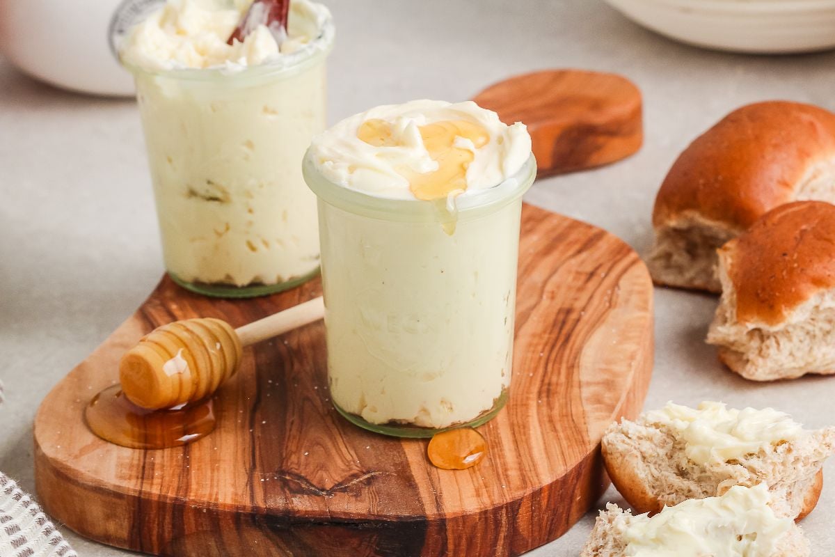 Two jars of homemade honey butter on a wooden board, with honey drizzled on top and a honey dipper nearby.