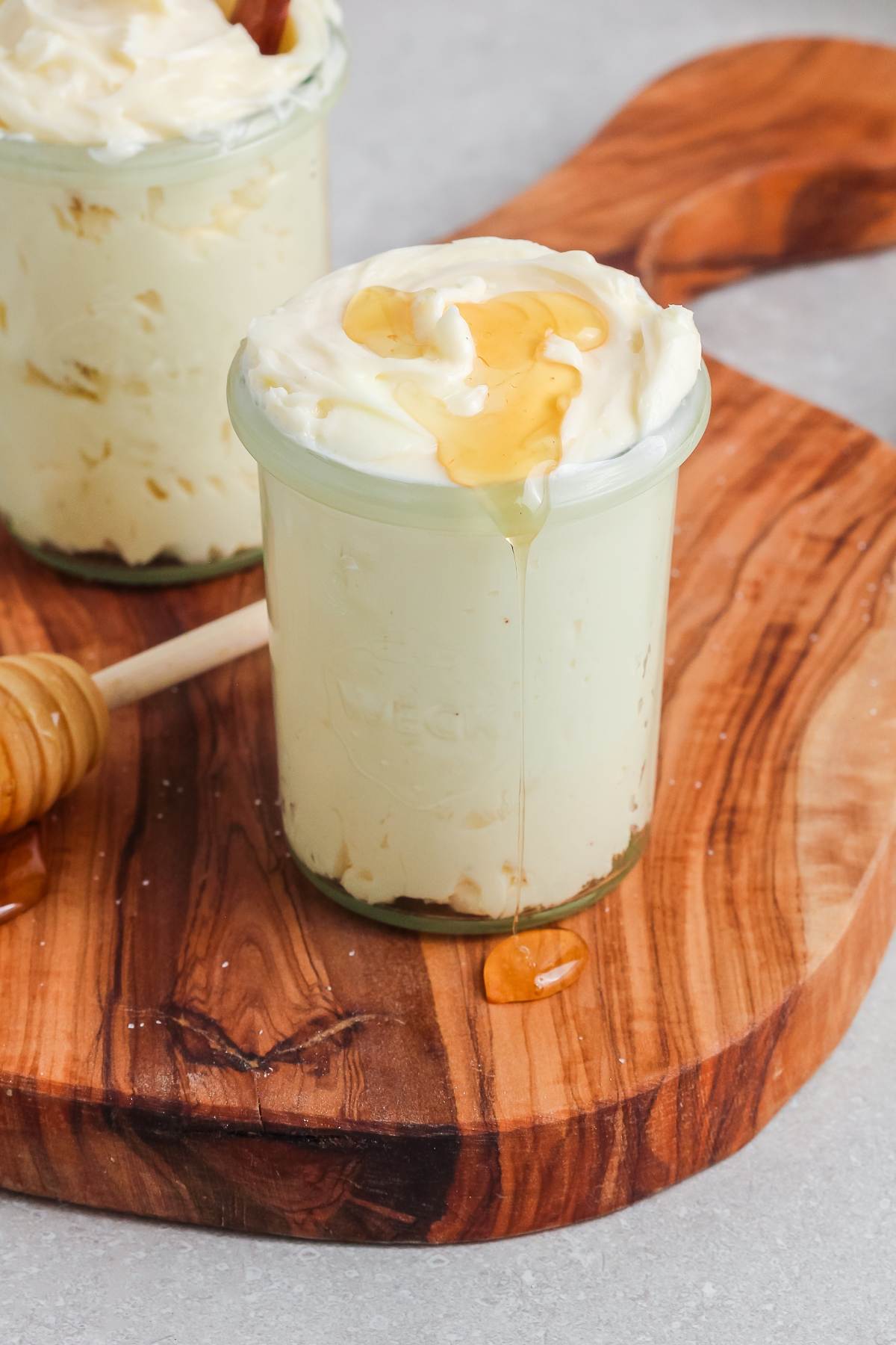 A glass jar of a butter spread topped with honey, sitting on a wooden board with a honey dipper nearby.