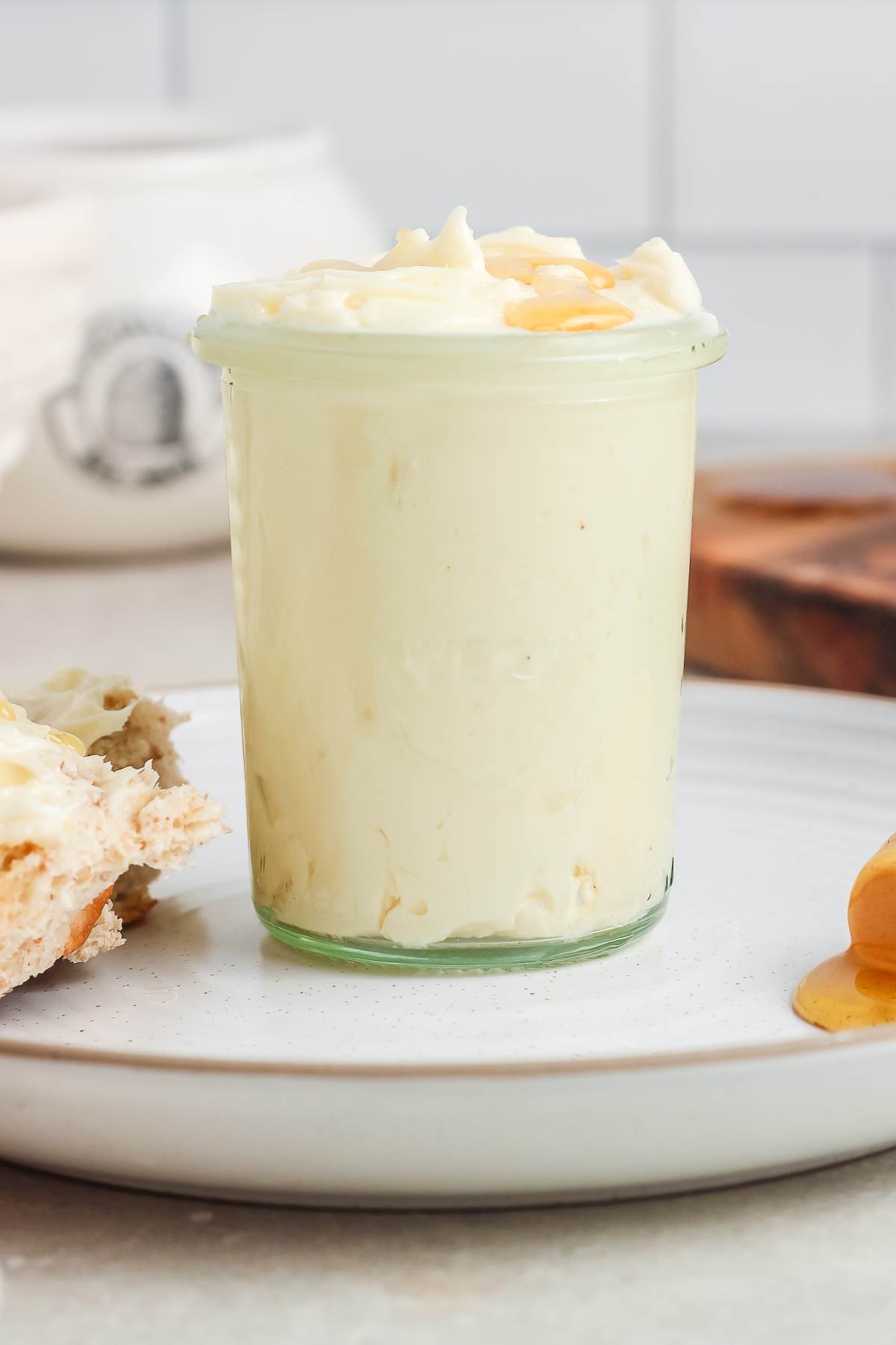 A glass jar filled with creamy honey butter sits on a white plate next to a slice of bread.