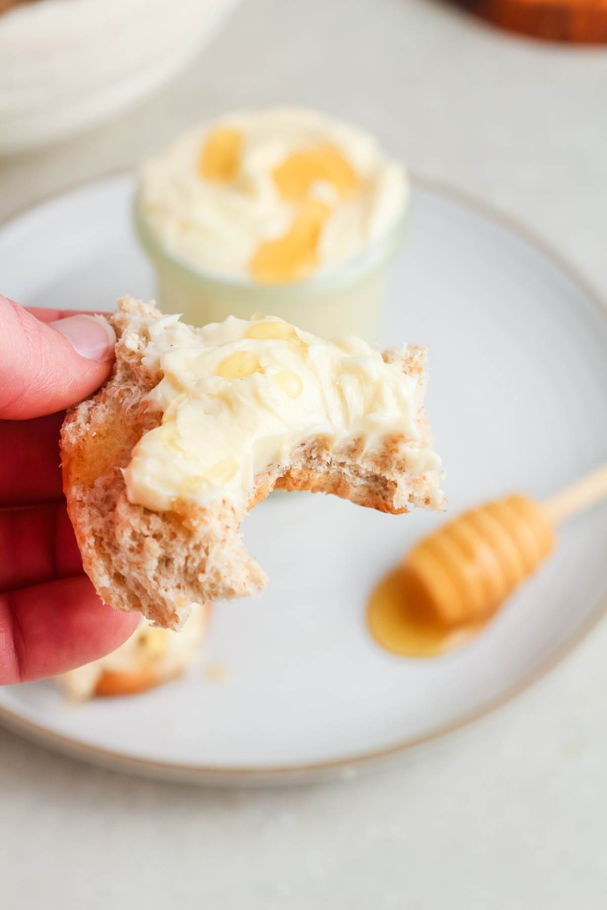 Hand holding bitten bread with honey butter, with honey dipper and more honey butter in background.