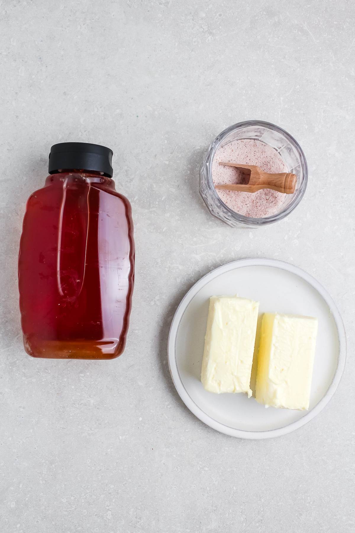 A bottle of honey, a bowl of pink salt, and two butter sticks on a small white plate.