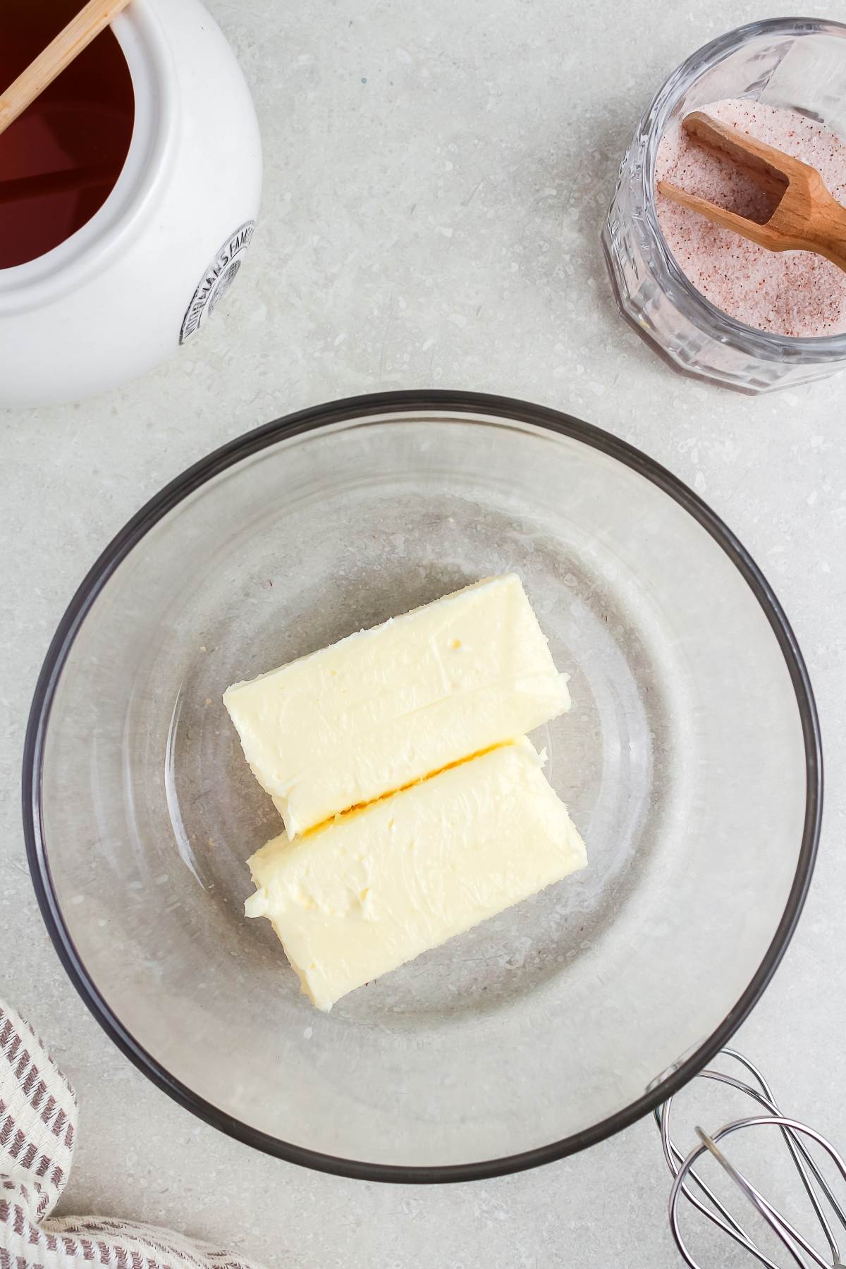 Two sticks of butter in a glass bowl next to a jar of honey and a small bowl of pink salt.