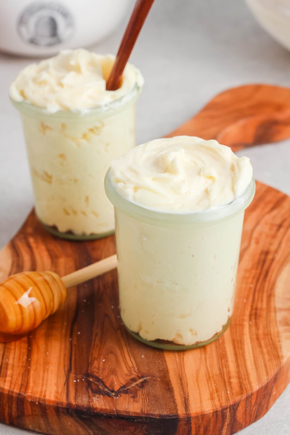 Two jars of whipped butter on a wooden board, with a honey dipper and a spoon in one jar.