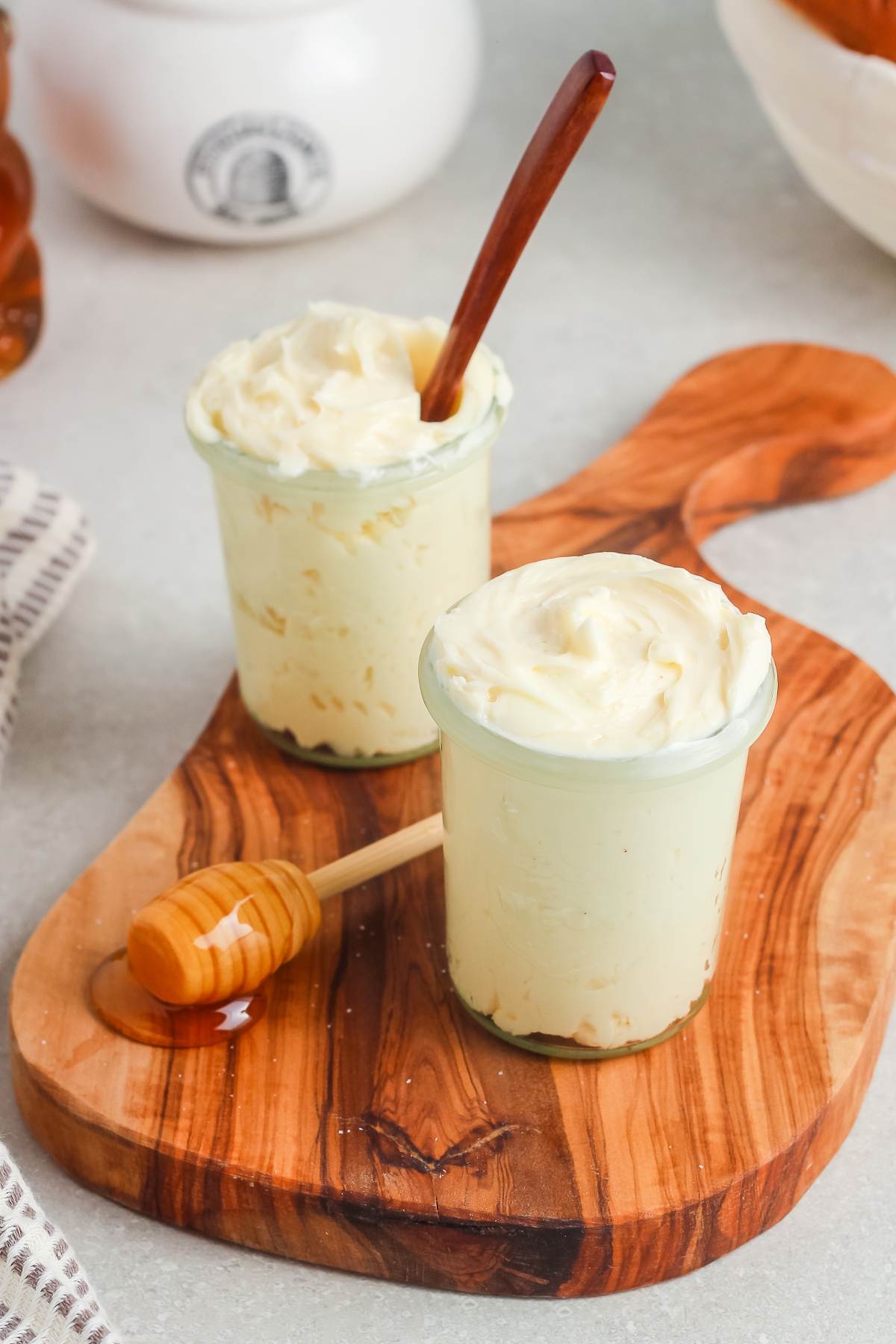 Two small glass jars of creamy butter on a wooden board, with a honey dipper nearby.