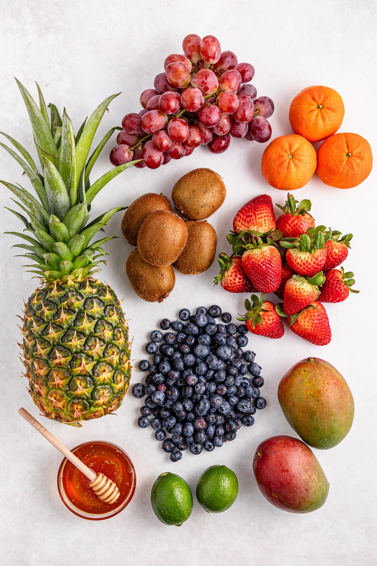 Assorted fresh fruits including pineapple, grapes, kiwis, strawberries, blueberries, mangoes, limes, mandarins, and honey.