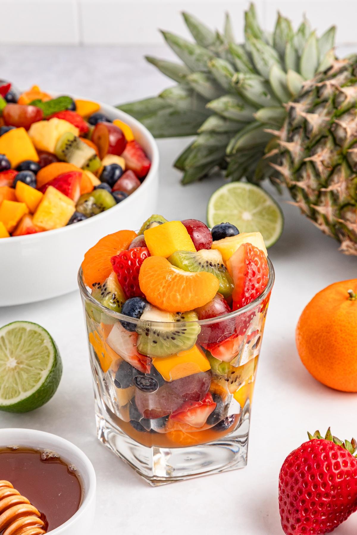 A glass filled with assorted fresh fruit salad, surrounded by whole fruits and a bowl of more fruit salad.