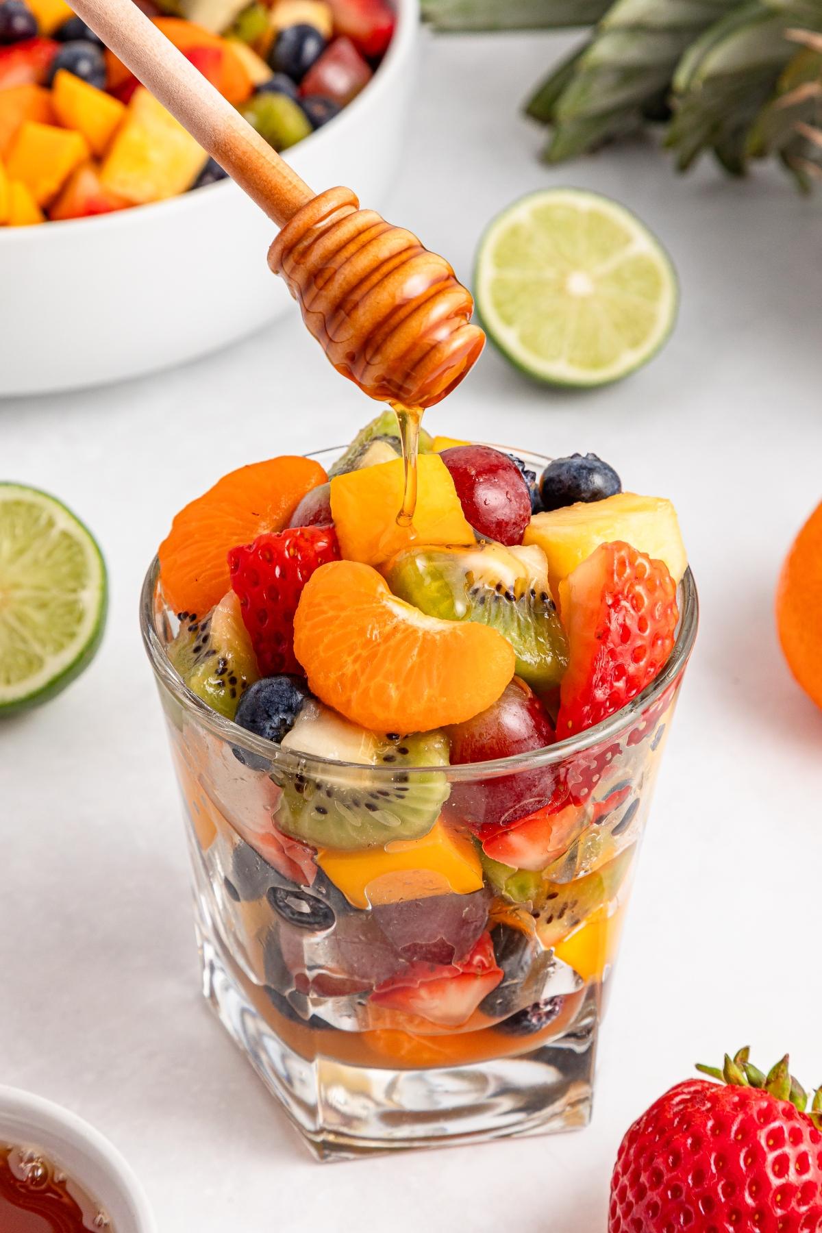 A glass of mixed fruit salad with honey being drizzled on top, surrounded by fresh fruits and lime halves.