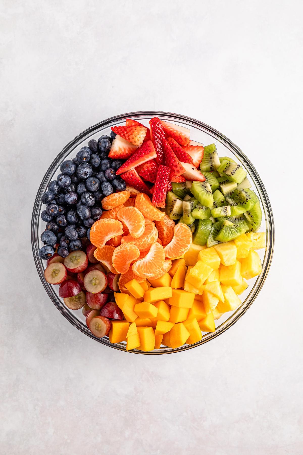 A glass bowl filled with strawberries, blueberries, kiwi, pineapple, grapes, oranges, and mango.