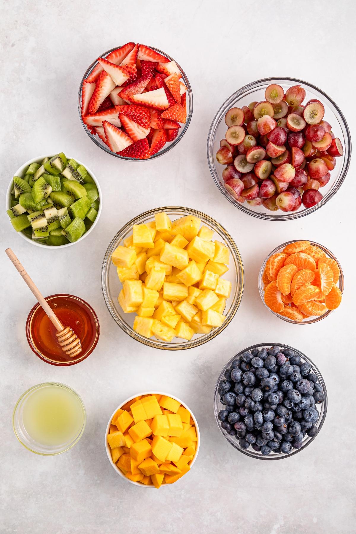 Bowls of sliced strawberries, grapes, kiwi, pineapple, mango, blueberries, mandarin, honey, and lemon juice.