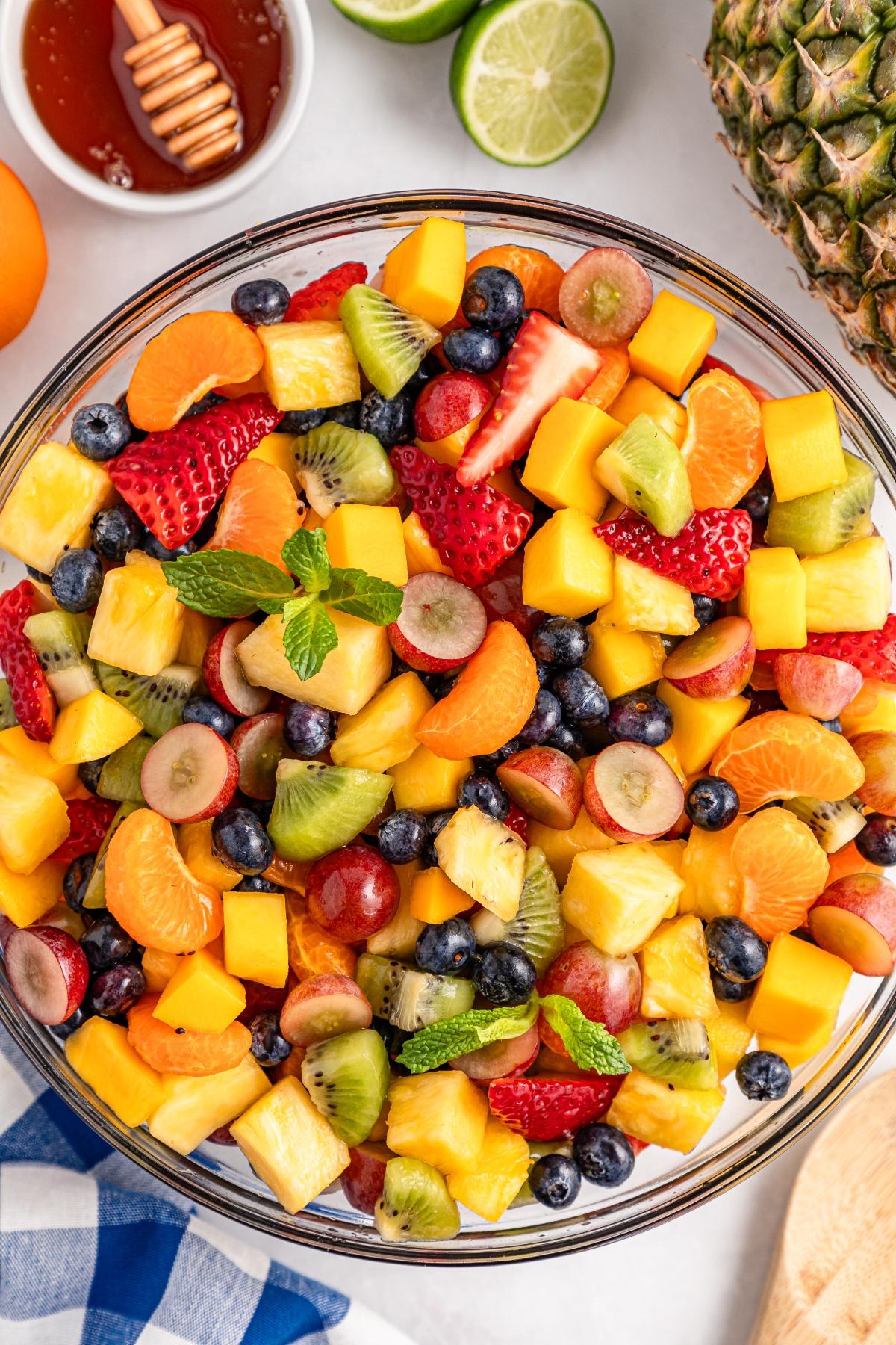 A bowl of colorful mixed fruit salad with strawberries, blueberries, kiwi, pineapple, grapes, and mint leaves.