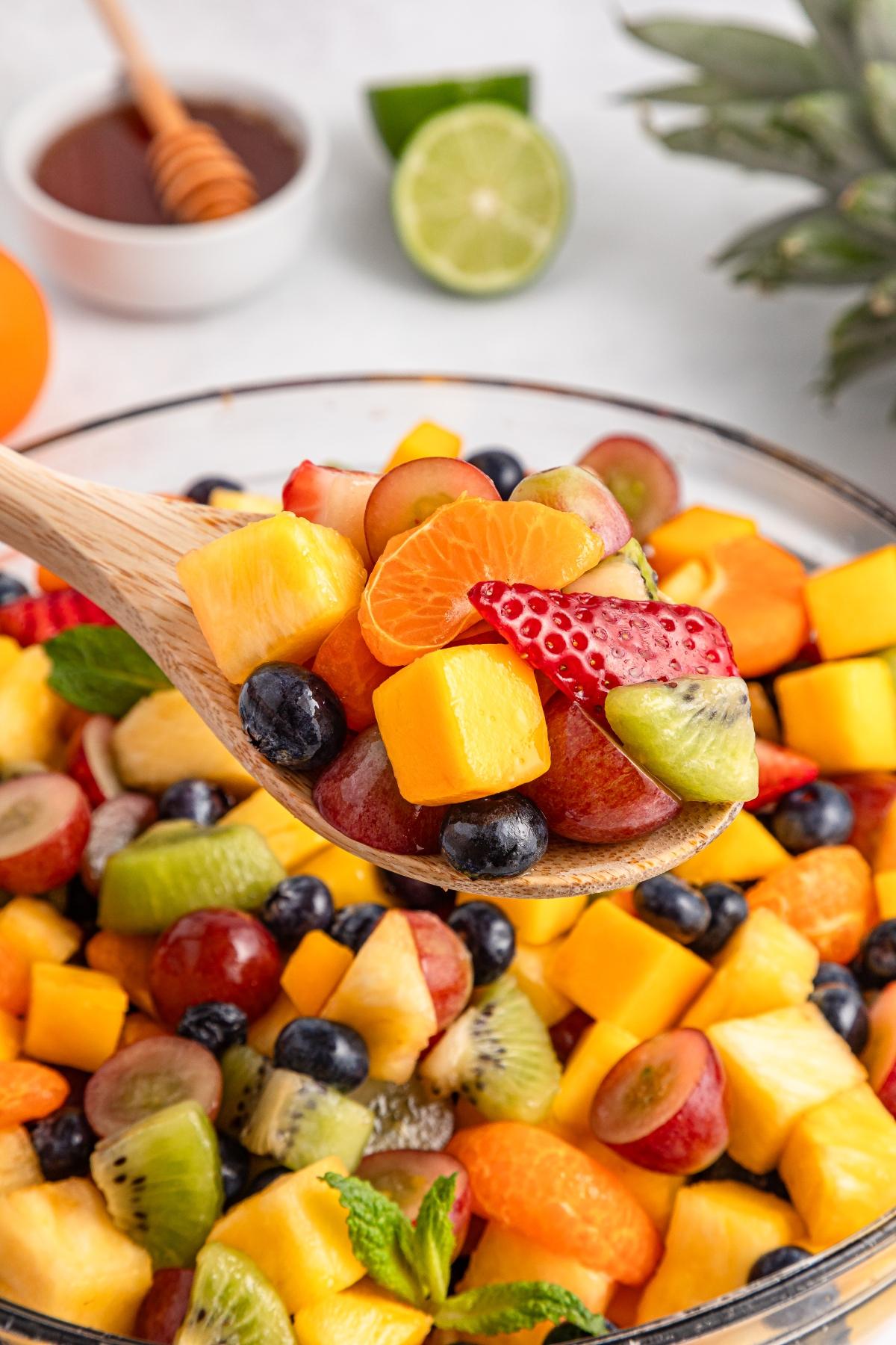 A wooden spoon holds assorted fresh fruit pieces above a colorful fruit salad bowl.
