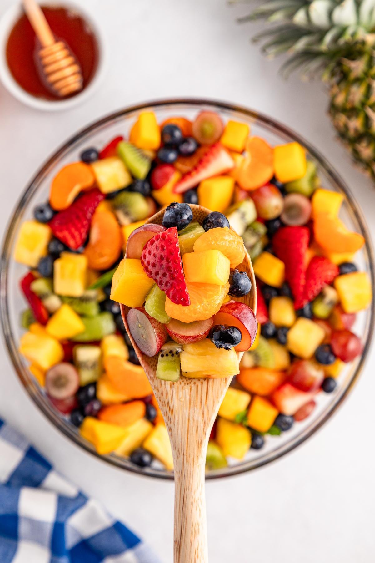 A wooden spoon holds colorful mixed fruit salad over a bowl, with honey and a pineapple nearby.