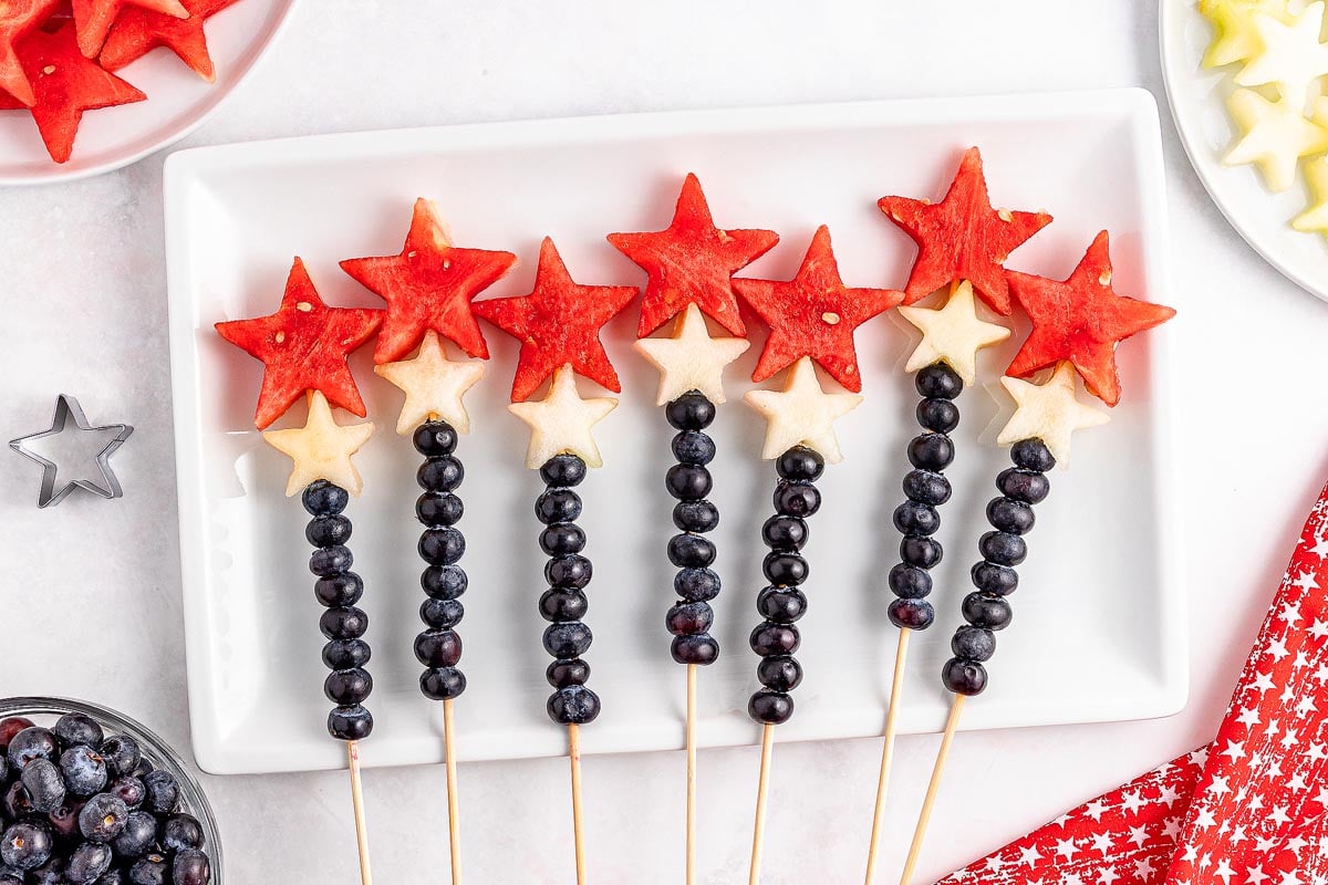 Fruit skewers featuring blueberries, white melon stars, and watermelon stars, beautifully arranged on a white rectangular plate.