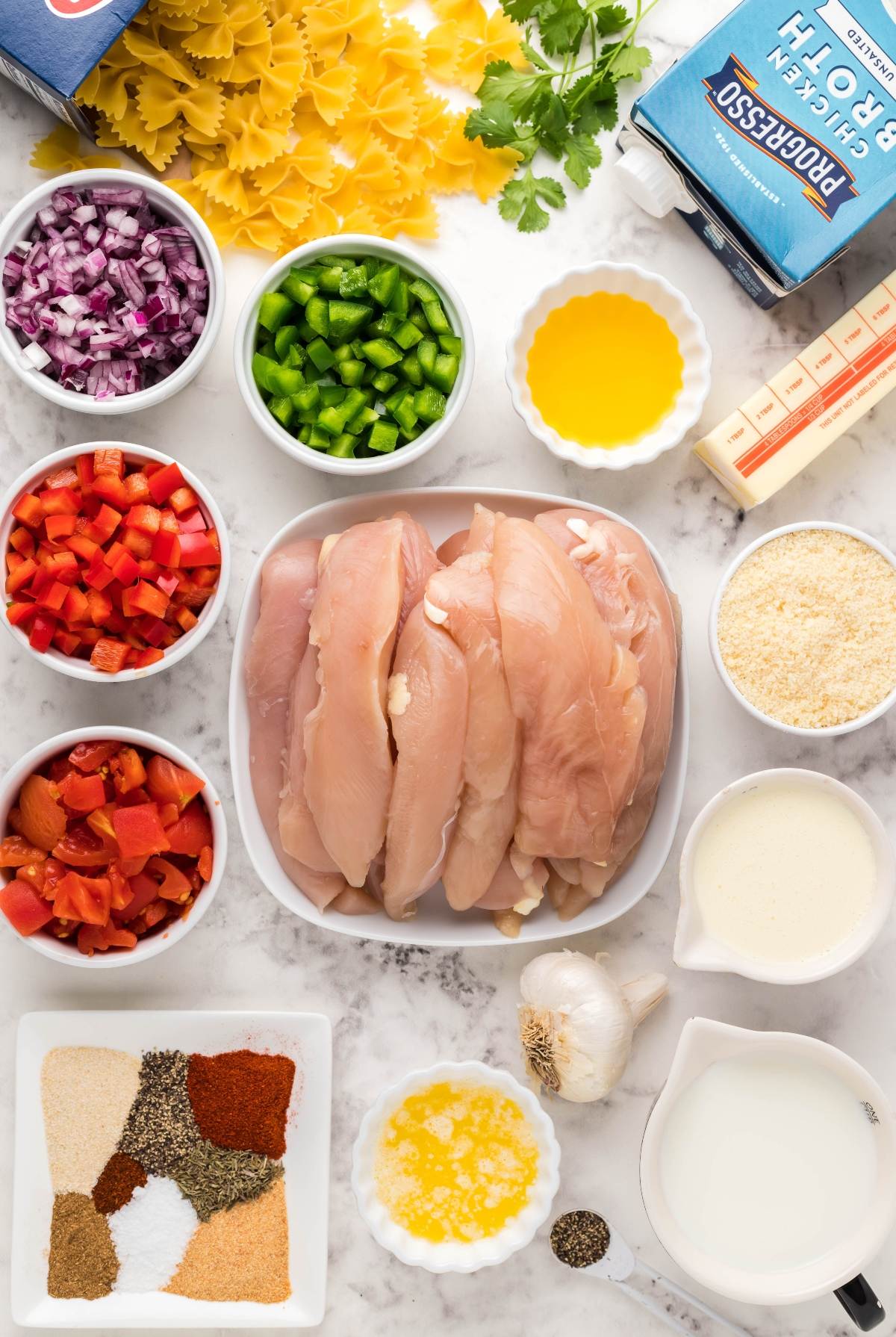 Raw chicken breasts surrounded by chopped vegetables, pasta, butter, broth, spices, cream, and cheese on a counter—everything you need for an easy instant pot chicken alfredo or quick instant pot chicken pasta dinner.