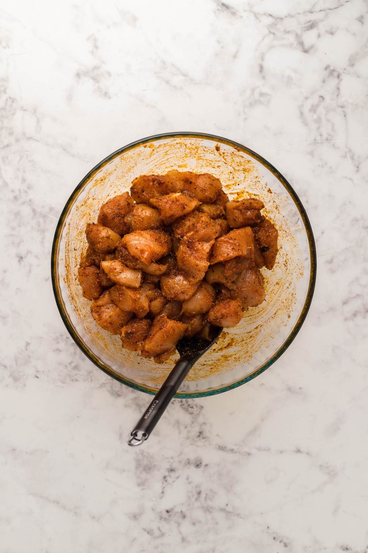 Glass bowl of raw, seasoned chicken pieces with a spoon, ready to be transformed into Instant Pot Blackened Chicken Alfredo on a white marble surface.