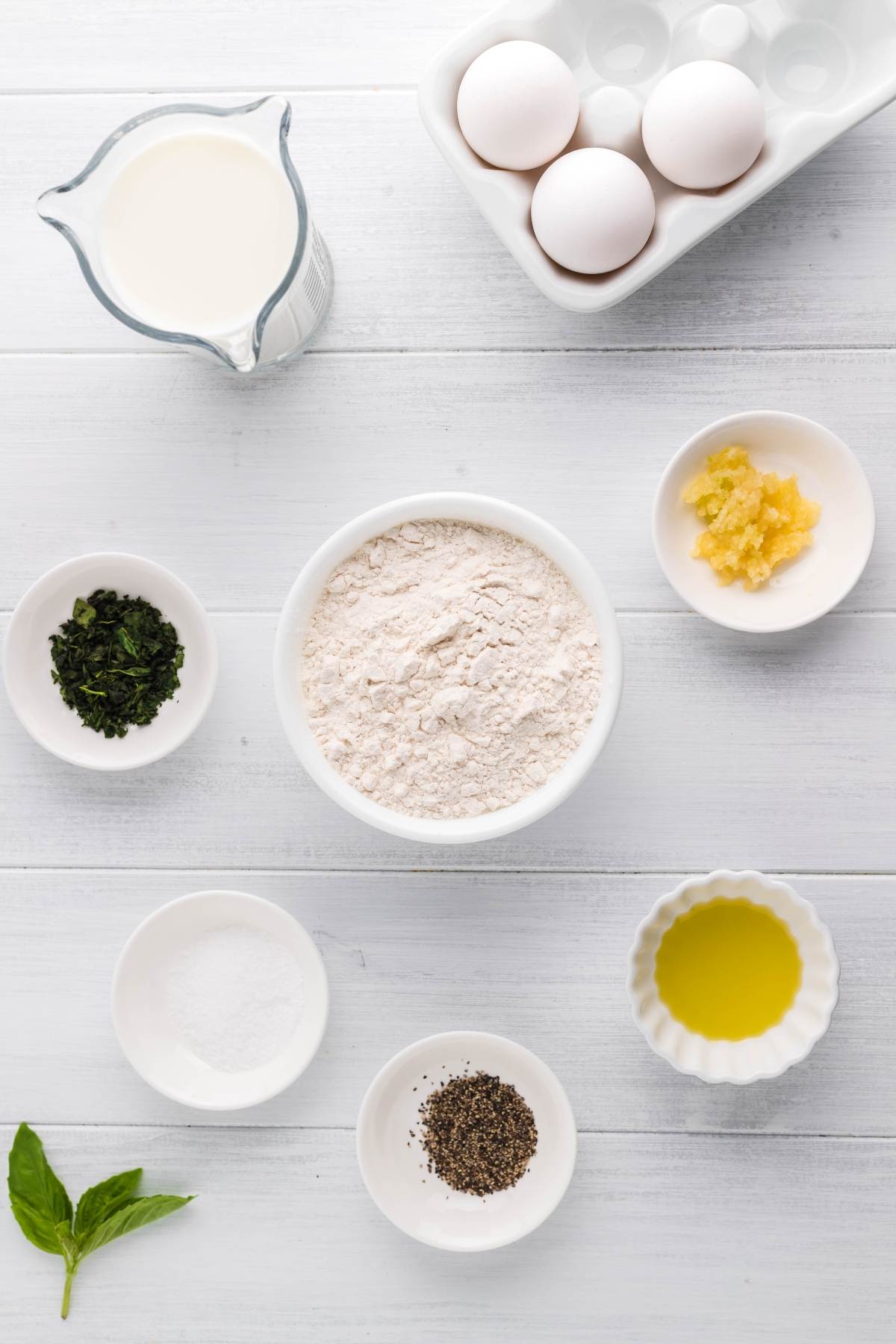 Ingredients on a white table for Italian crepes with Alfredo sauce: flour, eggs, milk, oil, herbs, minced garlic, pepper, salt, and a basil leaf.