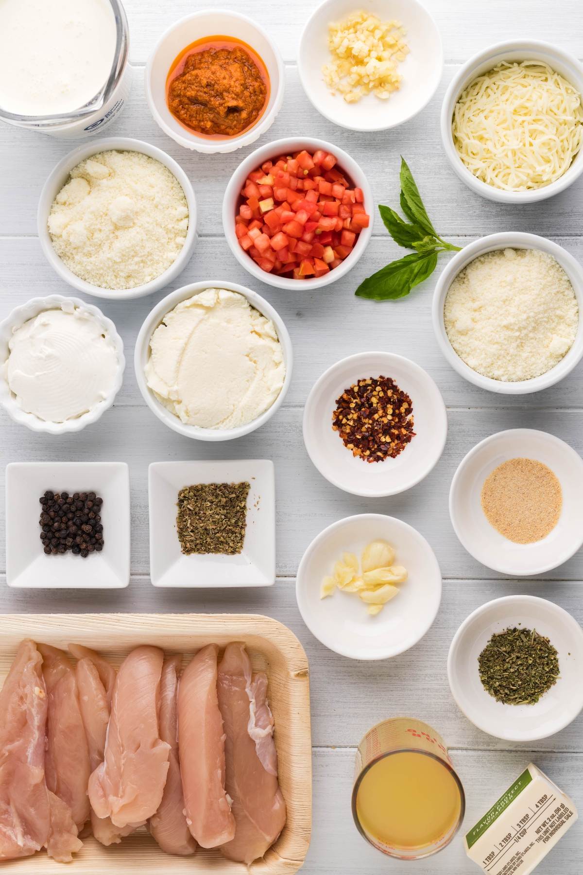 Raw chicken breasts, spices, cheeses, cream, diced peppers, herbs, garlic, and broth arranged on a white background—perfect for making Chicken Alfredo Crepes or using as a great leftover chicken recipe.