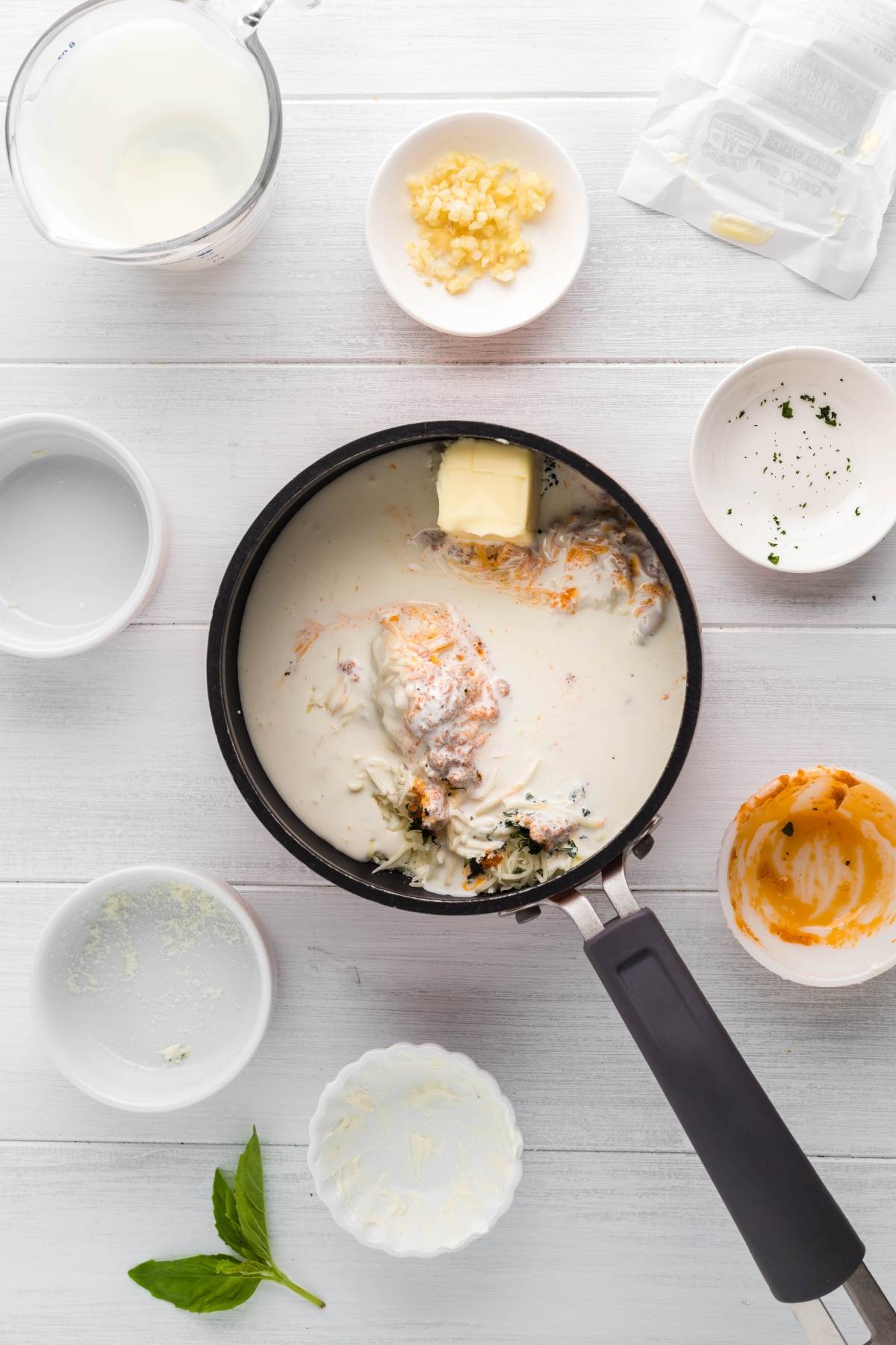 A saucepan with milk, butter, and seasonings, surrounded by bowls of ingredients on a white surface—perfect for making savoury chicken crepes or an Italian Crespelle - Chicken Alfredo Crepes with your leftover chicken.