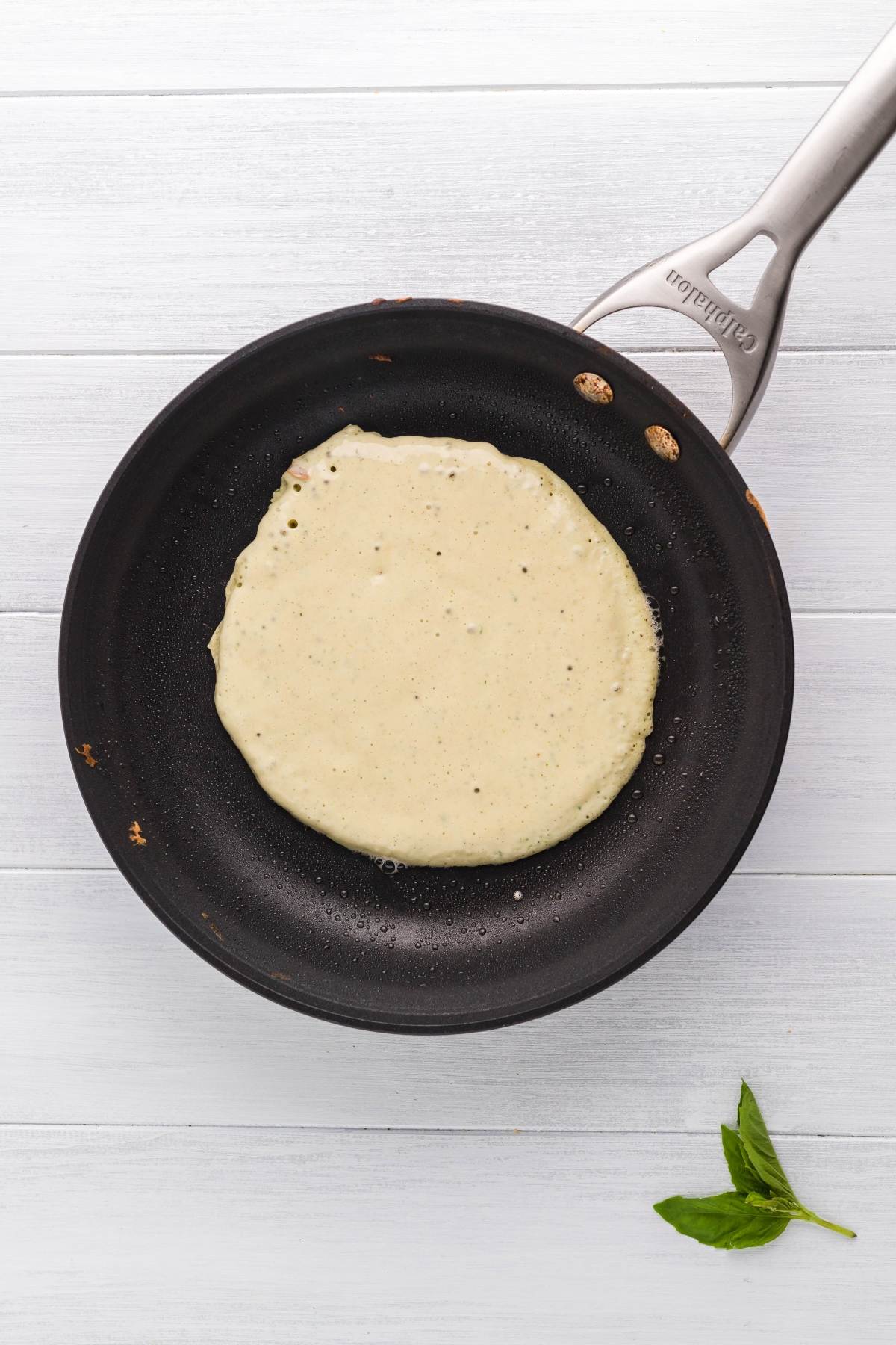 A pancake cooking in a black nonstick pan on a light wooden surface, with a basil leaf nearby—perfect inspiration for savoury chicken crepes.
