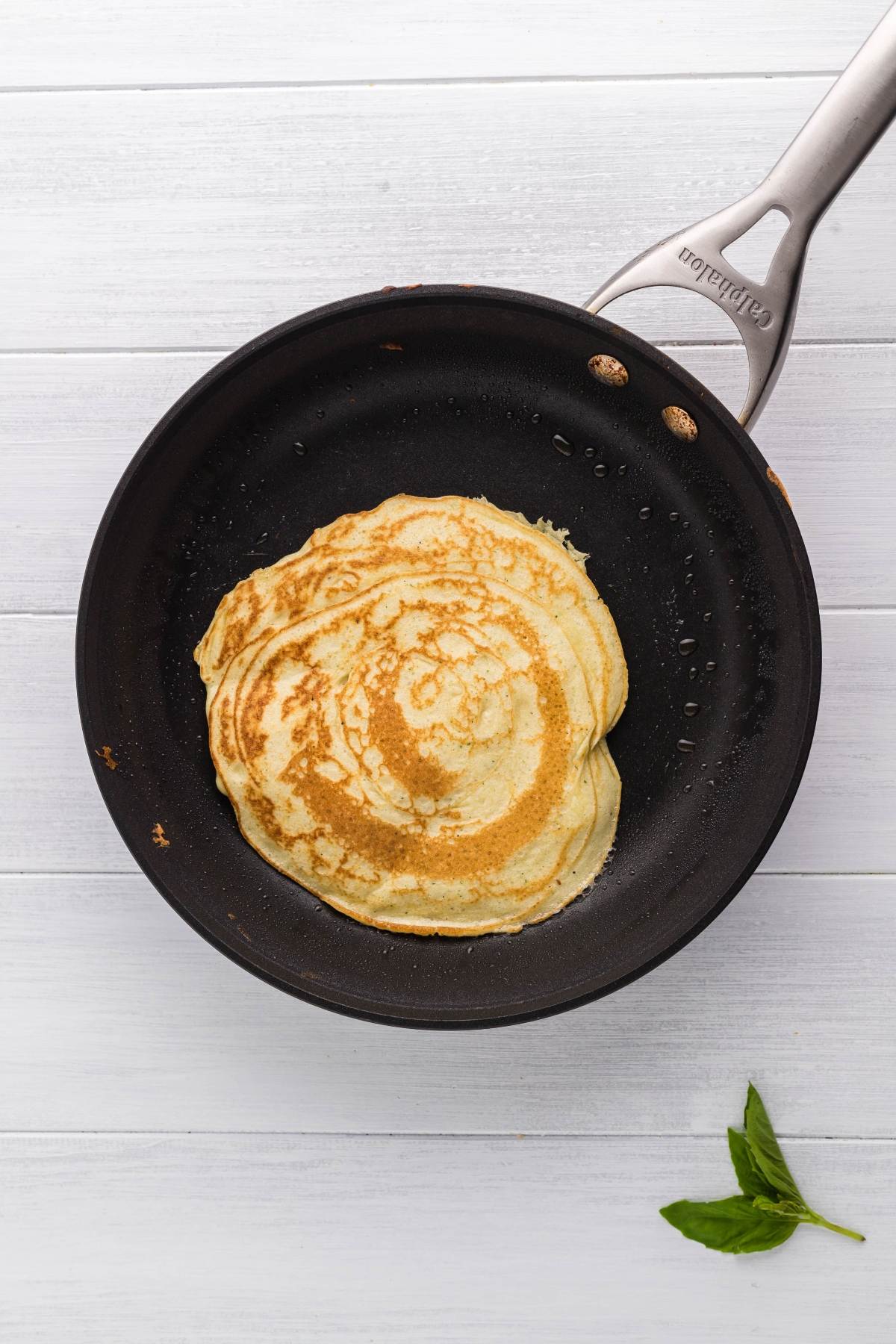 A pancake cooks in a black frying pan on a white wooden surface, with a basil leaf nearby—perfect inspiration for Italian Crespelle - Chicken Alfredo Crepes, a great leftover chicken recipe.