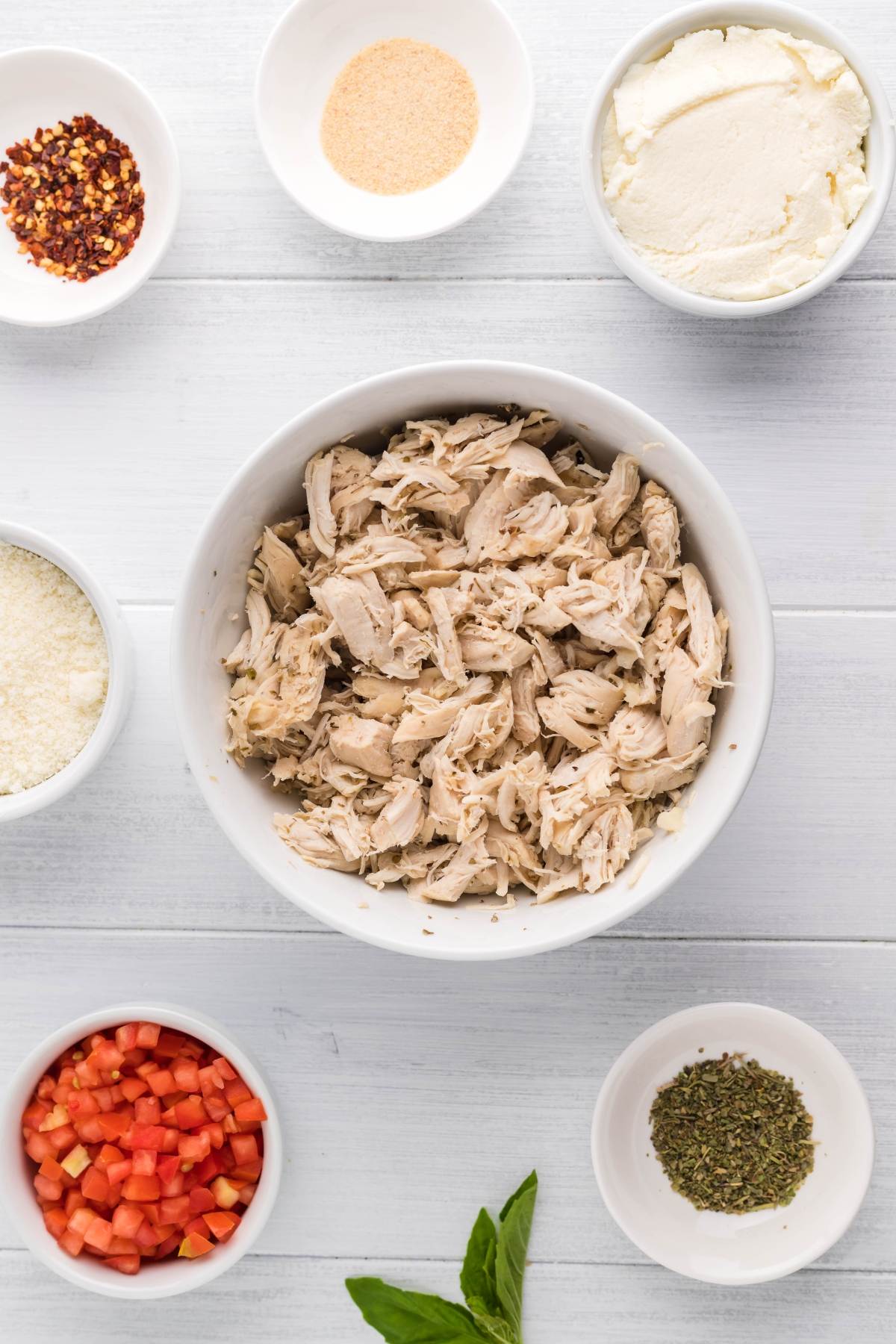 Overhead view of shredded chicken, cheese, diced peppers, and seasonings in white bowls on a white surface—perfect for making Chicken Alfredo Crepes, a great leftover chicken recipe.