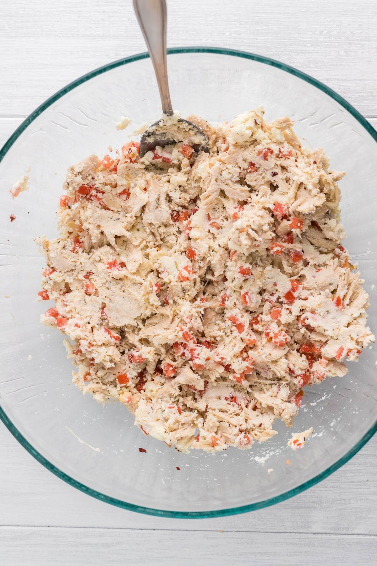 A glass bowl of shredded chicken mixed with chopped red peppers, perfect as a filling for savoury chicken crepes or delicious Italian crepes with Alfredo sauce. A spoon rests inside, ready to serve.