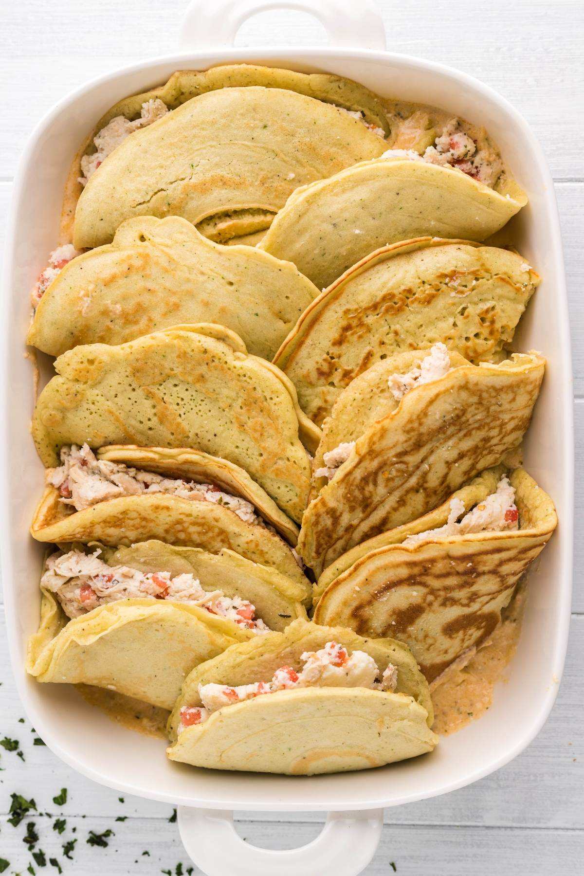 A white baking dish filled with Italian crepes with Alfredo sauce, stuffed and folded into savory crescents—Italian Crespelle - Chicken Alfredo Crepes are a great leftover chicken recipe for any light surface.