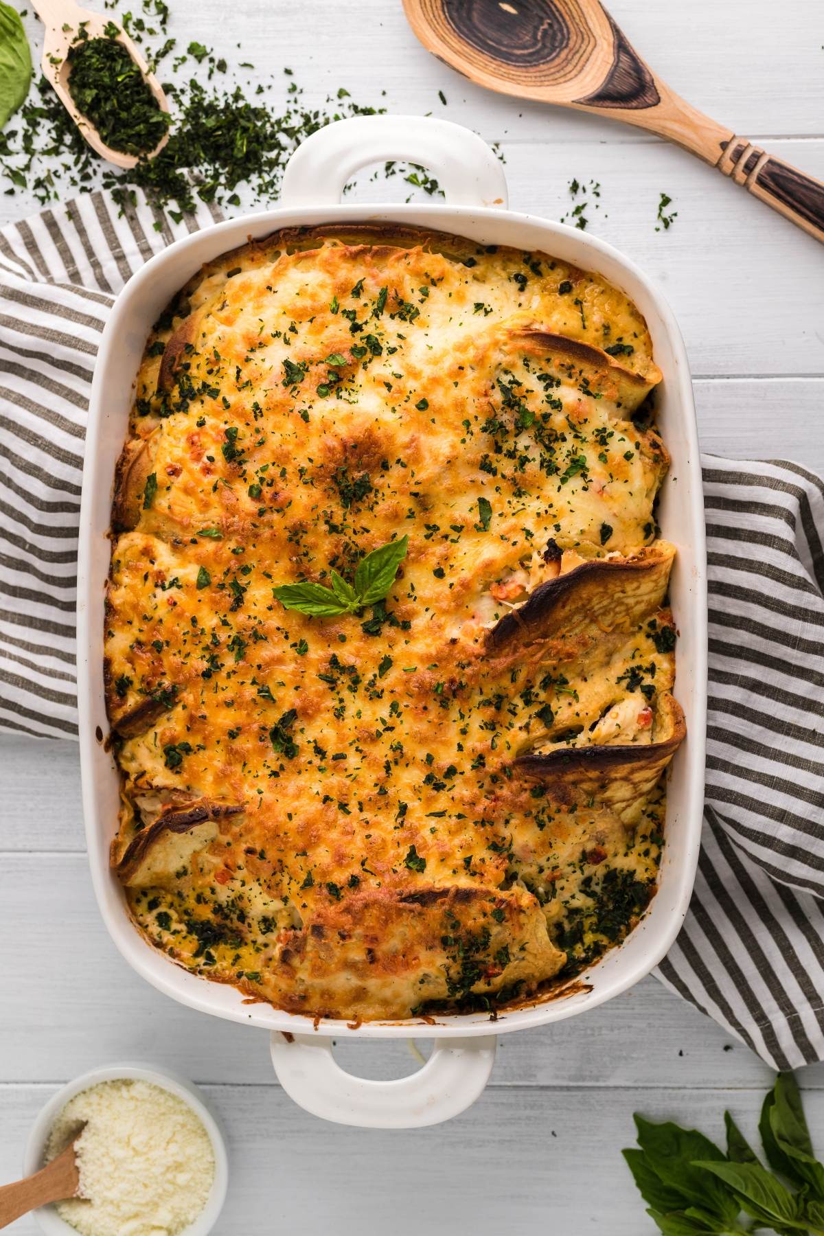Baked lasagna in a white dish, topped with melted cheese and herbs, shares the table with Italian Crespelle - Chicken Alfredo Crepes, all set on a striped cloth with utensils nearby.