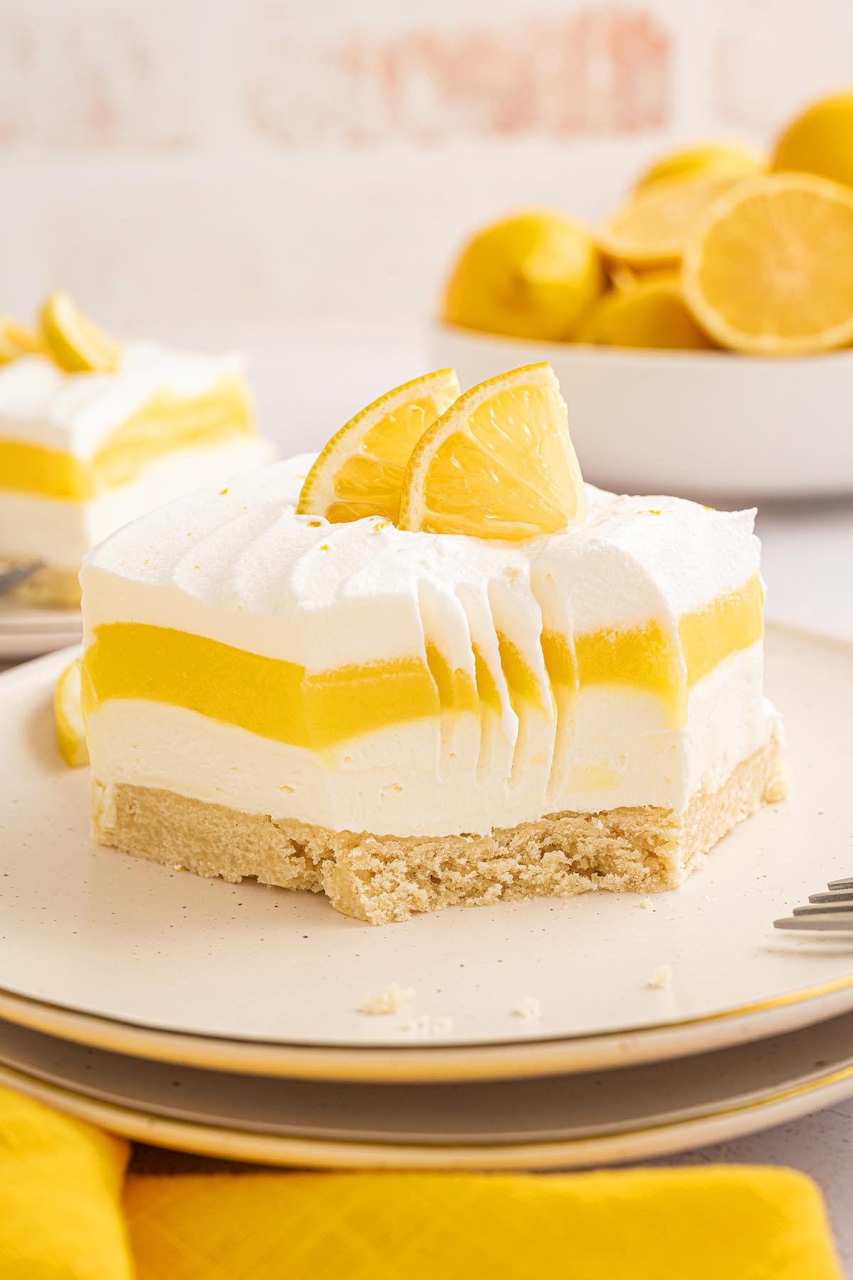 A slice of layered lemon lush dessert with whipped topping and lemon slices, with a bite taken out, on a plate.
