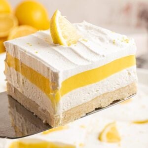A slice of lemon lush dessert with whipped topping and a lemon wedge, held on a spatula.