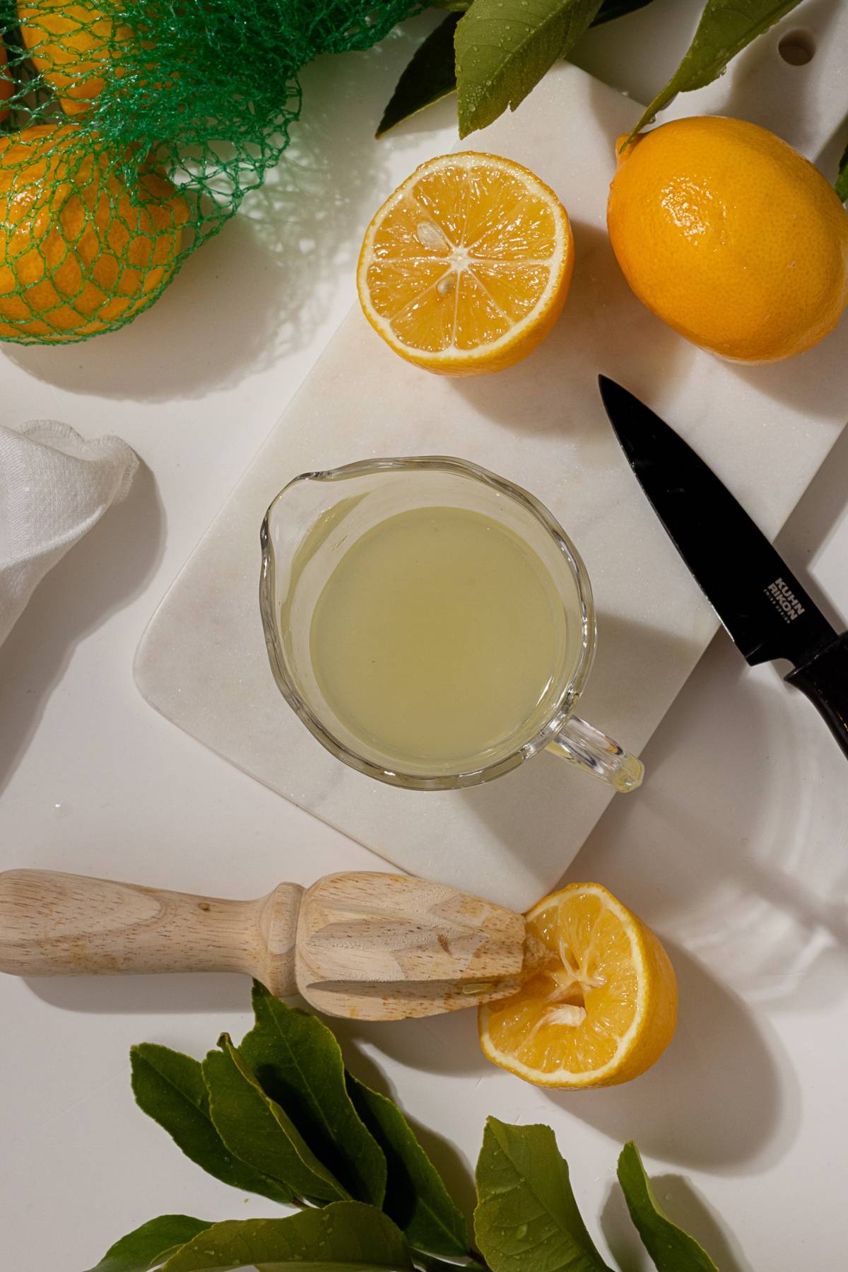 Glass cup of lemon juice, cut lemons, leaves, a knife, and wooden reamer on a white cutting board.