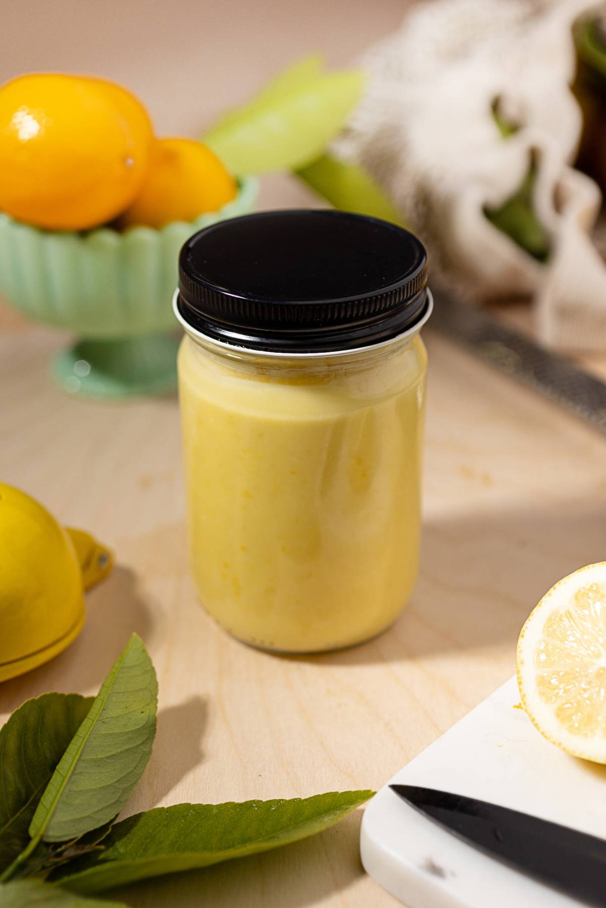 A jar of yellow lemon curd on a wooden surface, with fresh lemons and leaves nearby.