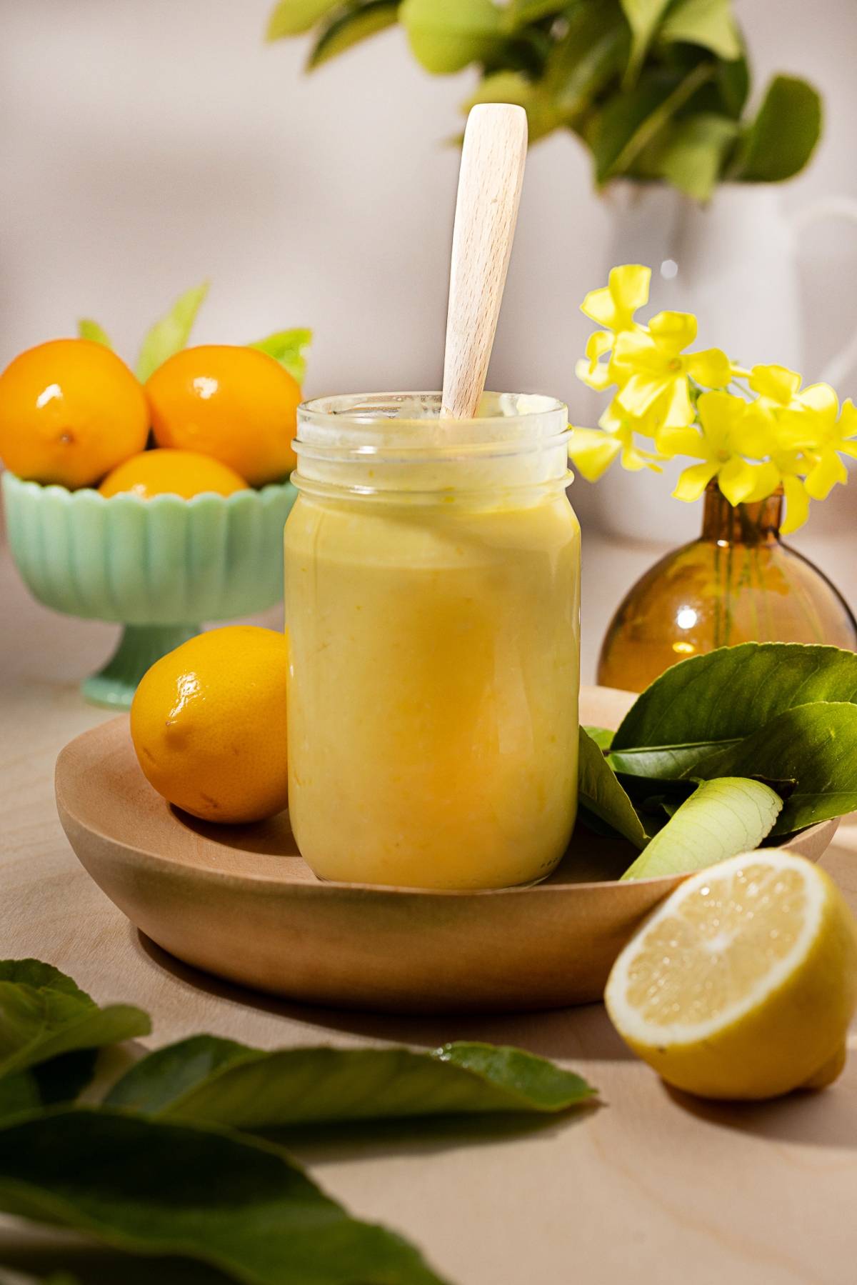 A jar of lemon curd with a spoon, surrounded by fresh lemons and green leaves on a wooden table.
