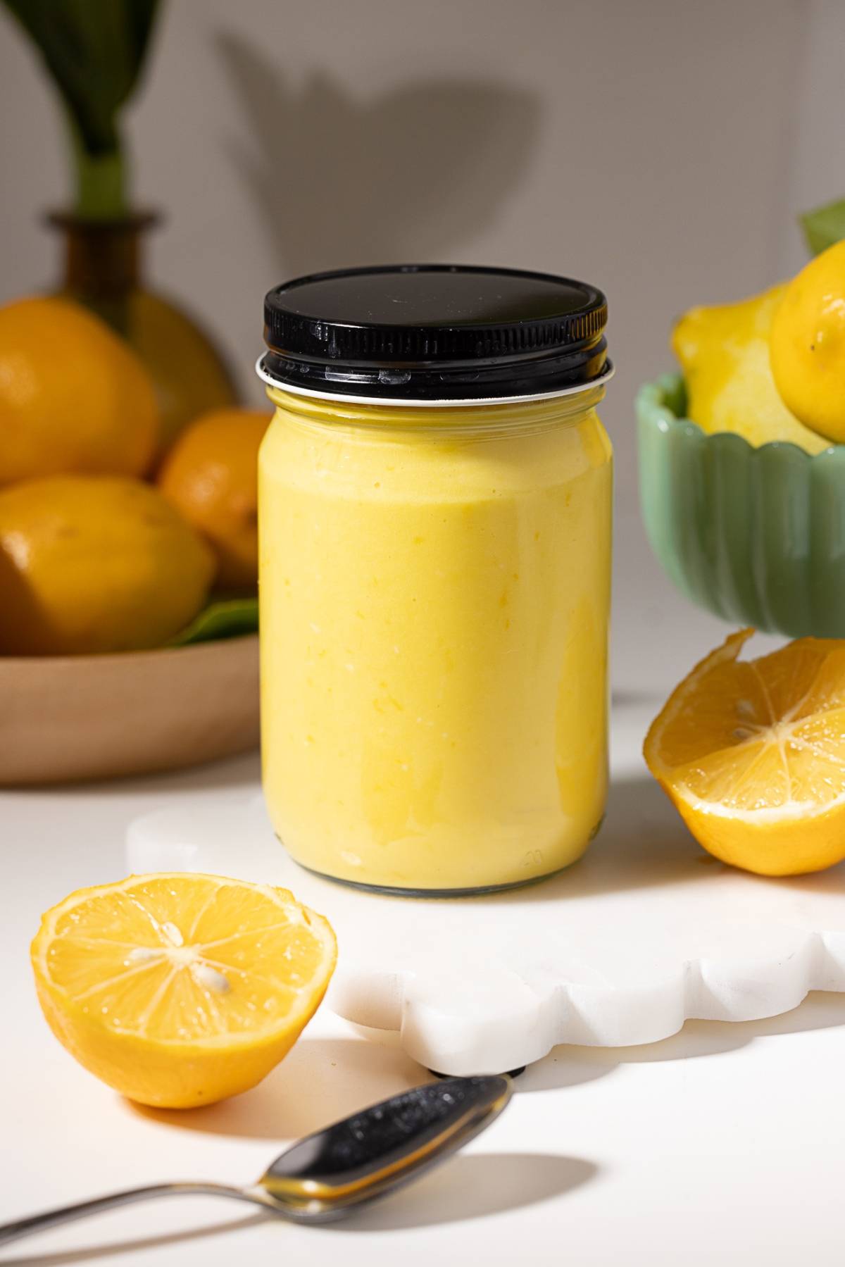 A jar of yellow lemon curd surrounded by fresh lemons and lemon halves on a white surface.