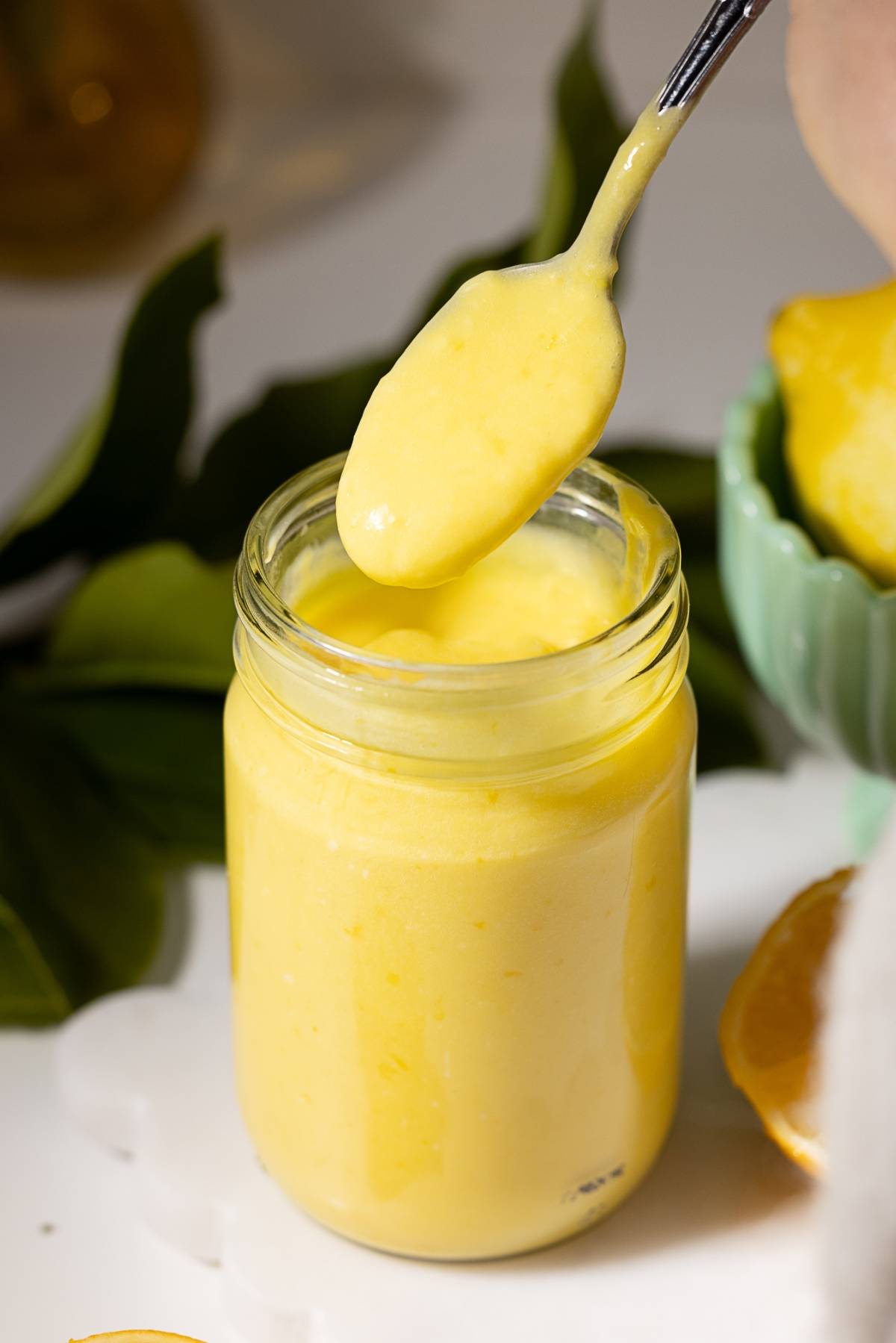A spoon scooping creamy yellow lemon curd from a glass jar, with lemons in the background.