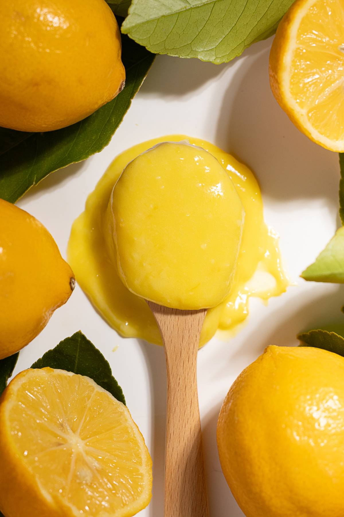A wooden spoon with lemon curd, surrounded by fresh lemons and green leaves on a white surface.