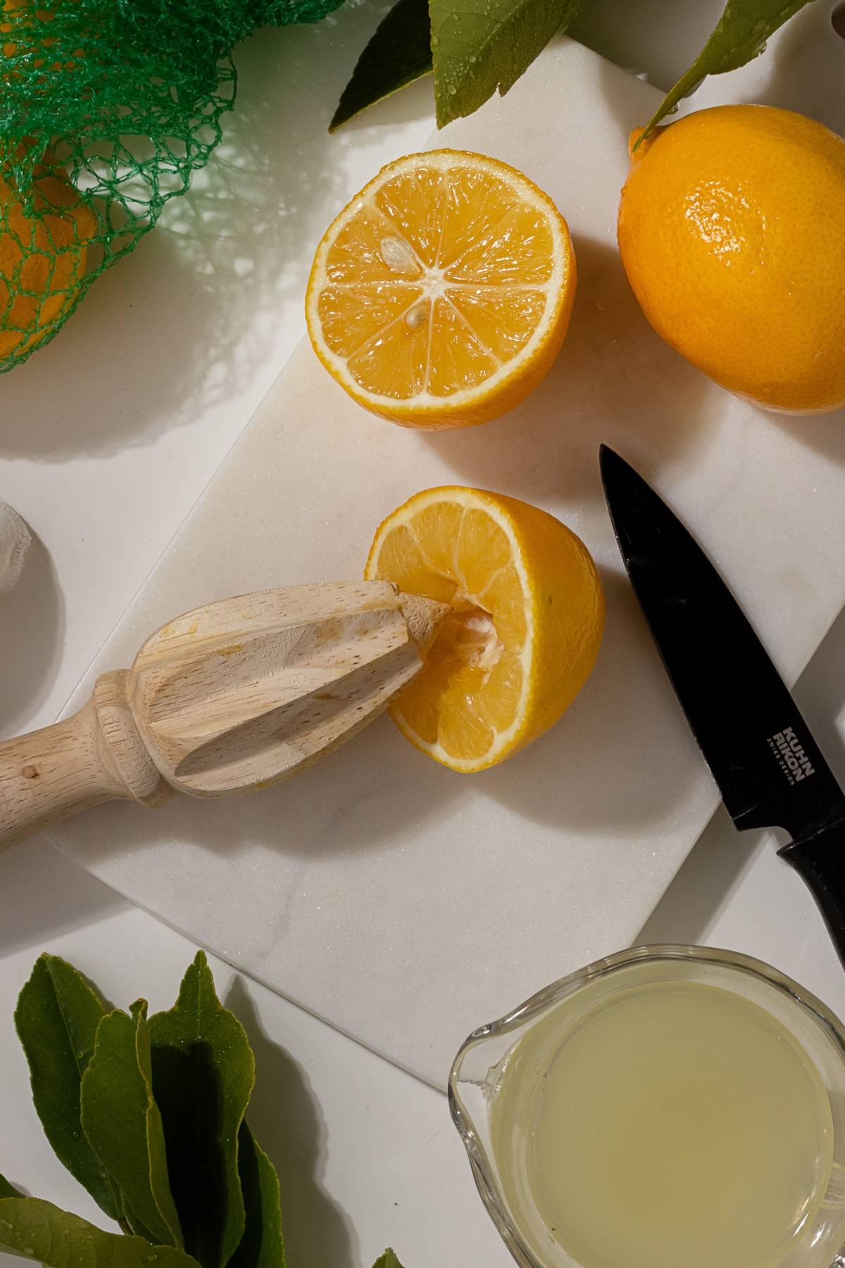 Sliced lemons, a juicer, a knife, and a cup of lemon juice on a white cutting board.