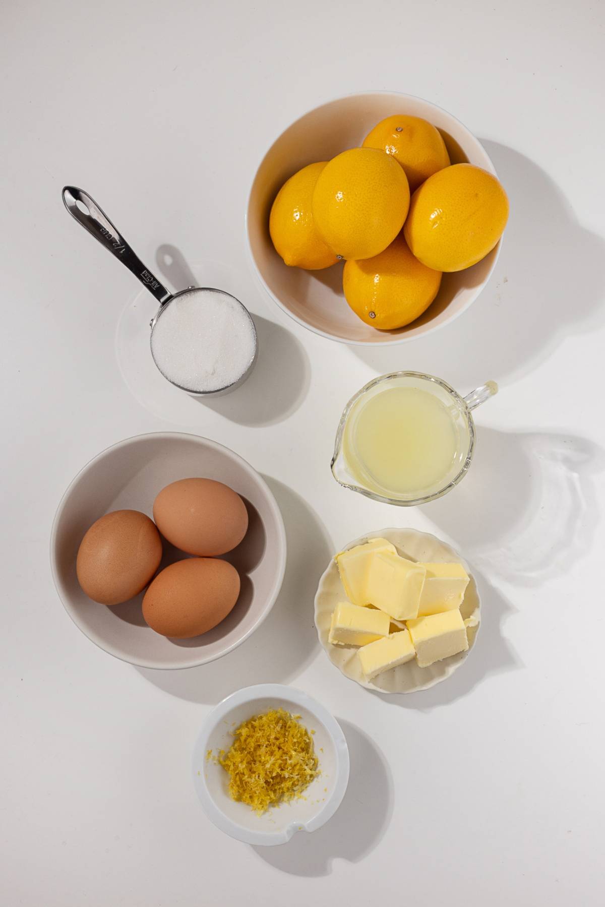Ingredients on a white surface: lemons, eggs, sugar, butter, lemon zest, and lemon juice in bowls and cups.