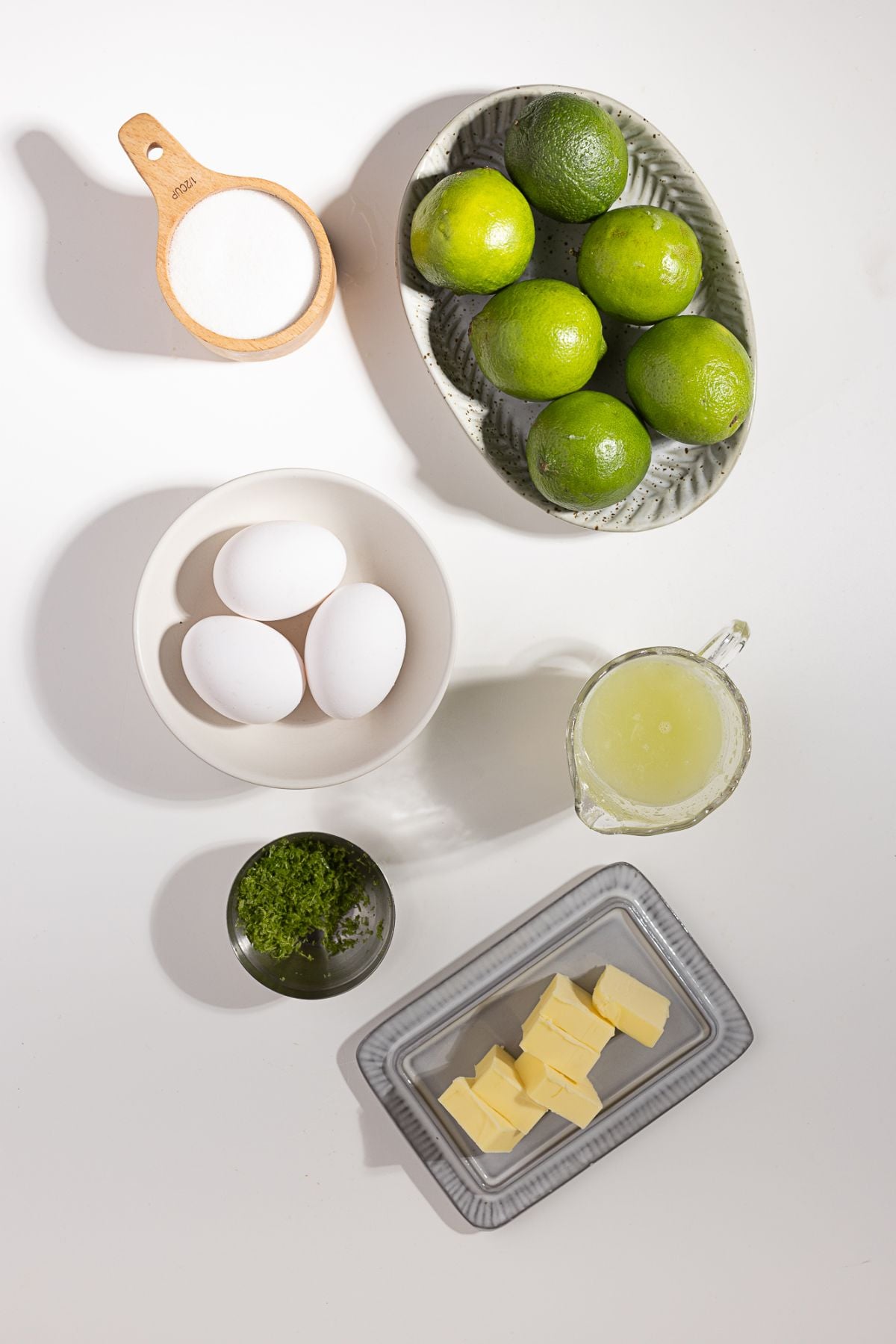 Limes, eggs, sugar, butter, lime zest, and lime juice arranged on a white surface—everything you need to make delicious homemade lime curd.