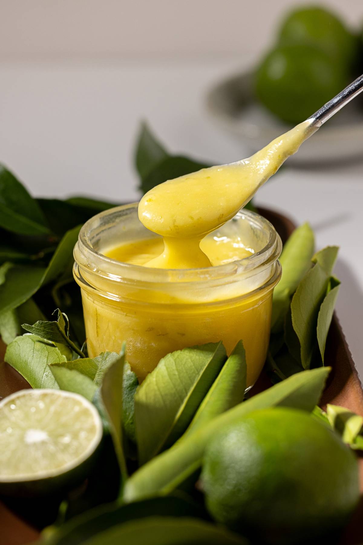 A spoonful of creamy yellow curd being lifted from a jar, surrounded by fresh limes and green leaves.